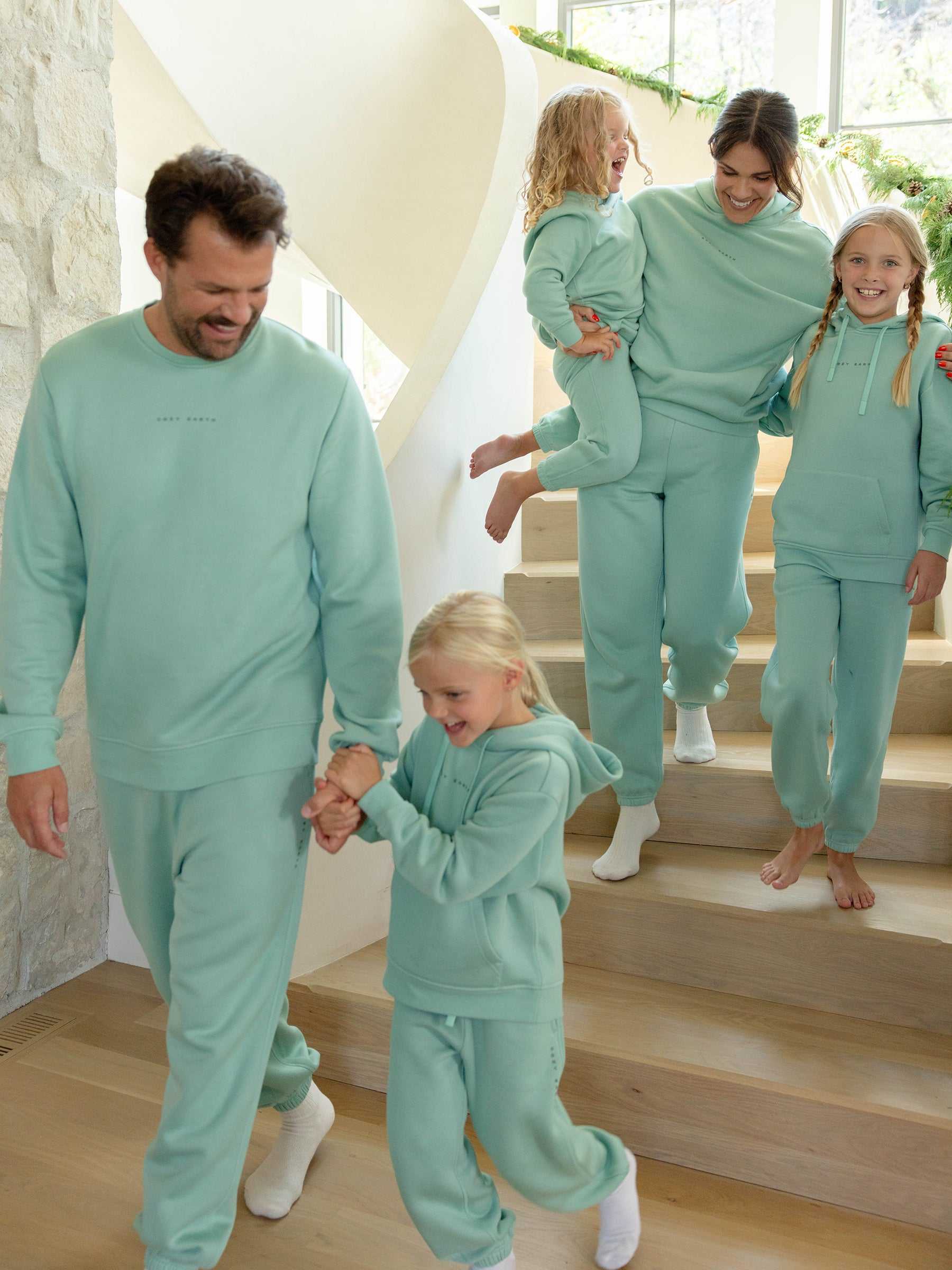 A family of five, all wearing matching Men's CityScape Crewnecks from Cozy Earth, cheerfully descends a wooden staircase. One adult carries a young child, while the others walk hand in hand. The space is filled with bright, natural light. |Color:Wintergreen