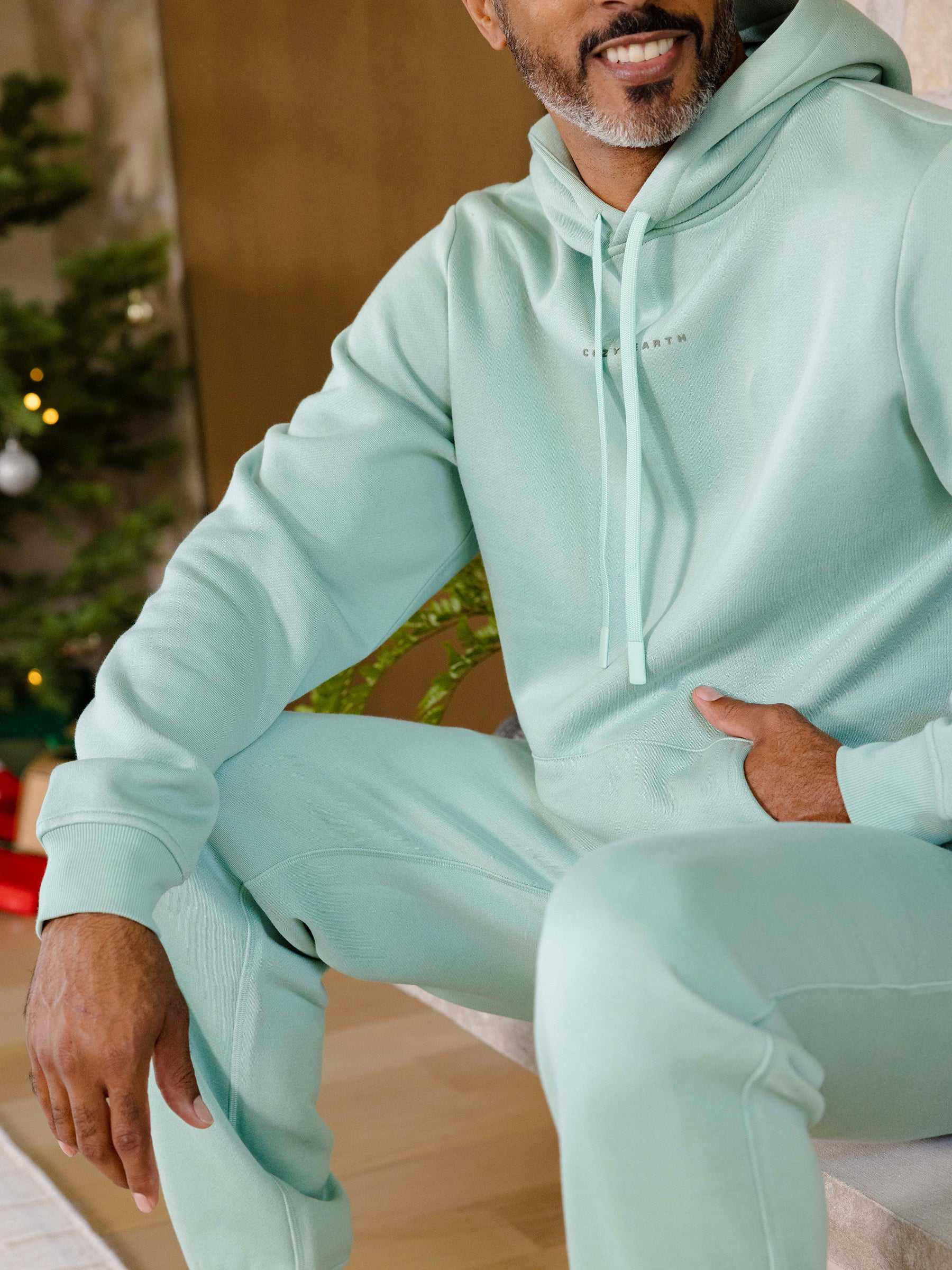 A person in a mint green Men's CityScape Hoodie and matching sweatpants from Cozy Earth sits casually. A Christmas tree with lights can be seen in the background, creating a festive atmosphere. |Color:Wintergreen