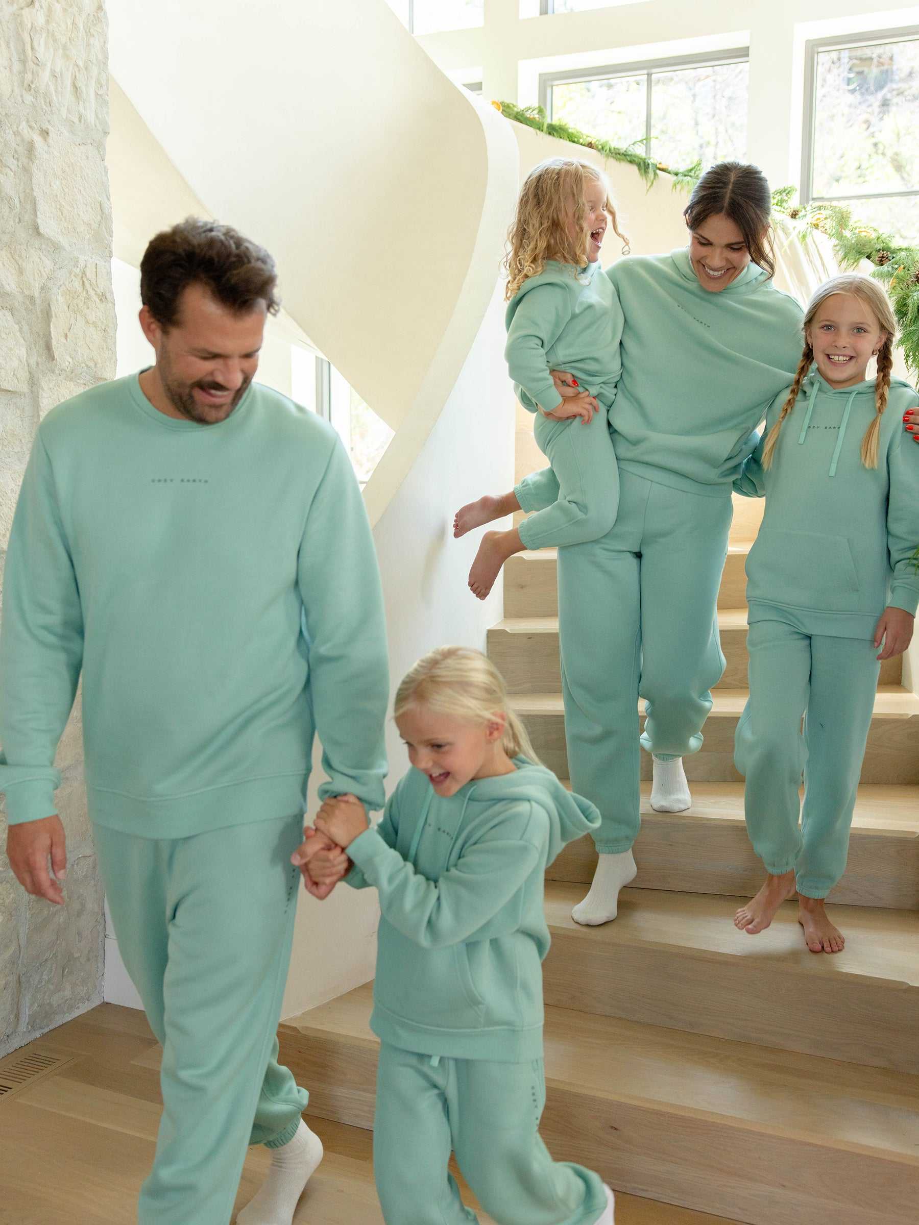 A family of five, clad in matching turquoise CityScape tracksuits by Cozy Earth, joyfully descends a modern staircase. The father, wearing the Men's CityScape Sweatpant, holds hands with one child at the front, while the mother carries another child and walks alongside their third child who is smiling brightly beside her. |Color:Wintergreen