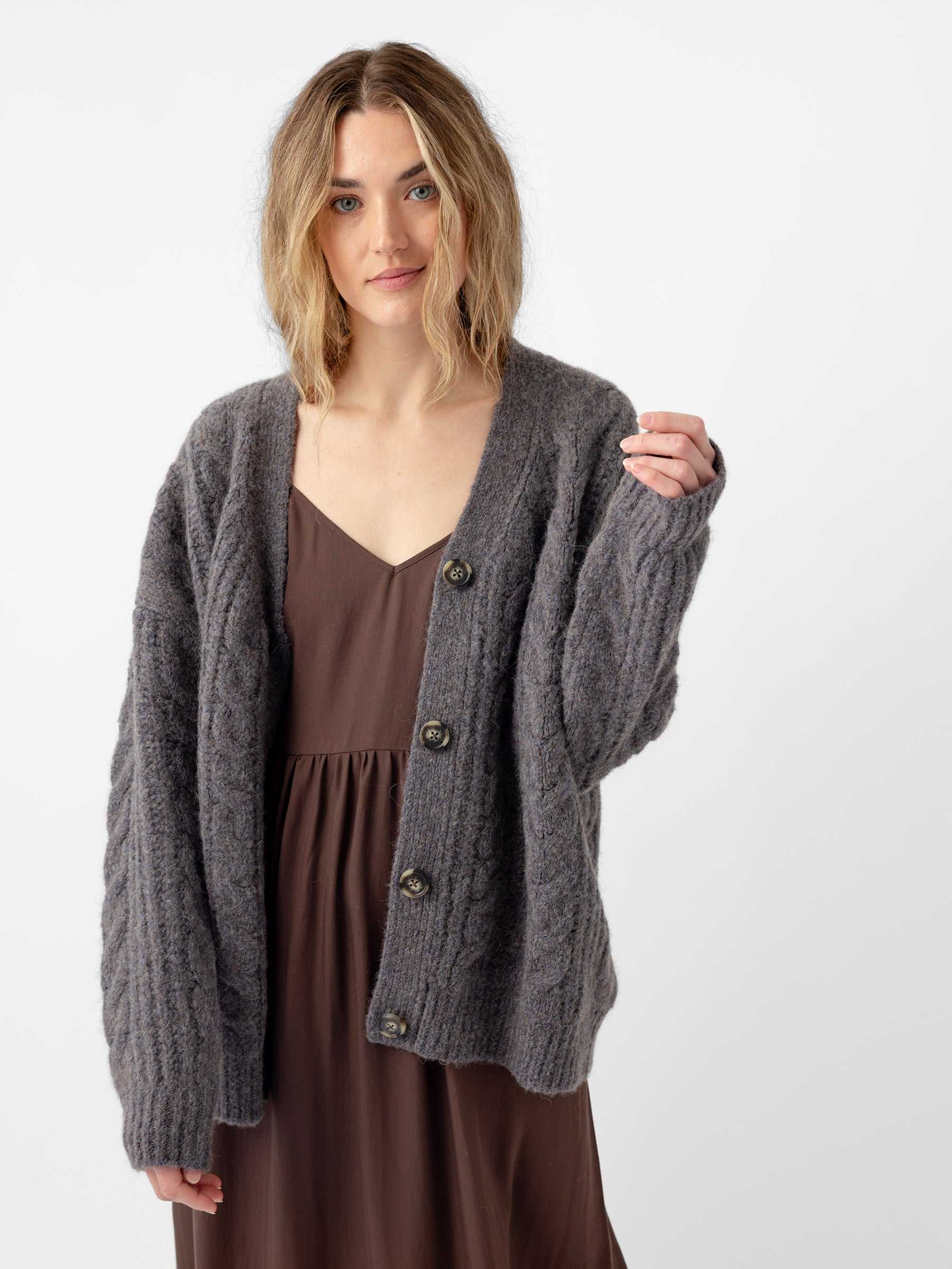 A person with wavy hair is wearing a dark brown dress and a Cozy Earth Oversized Cable Knit Cardigan in gray, featuring buttons. They stand against a plain white background, with a relaxed expression and slightly raised hand. |Color:Thunder