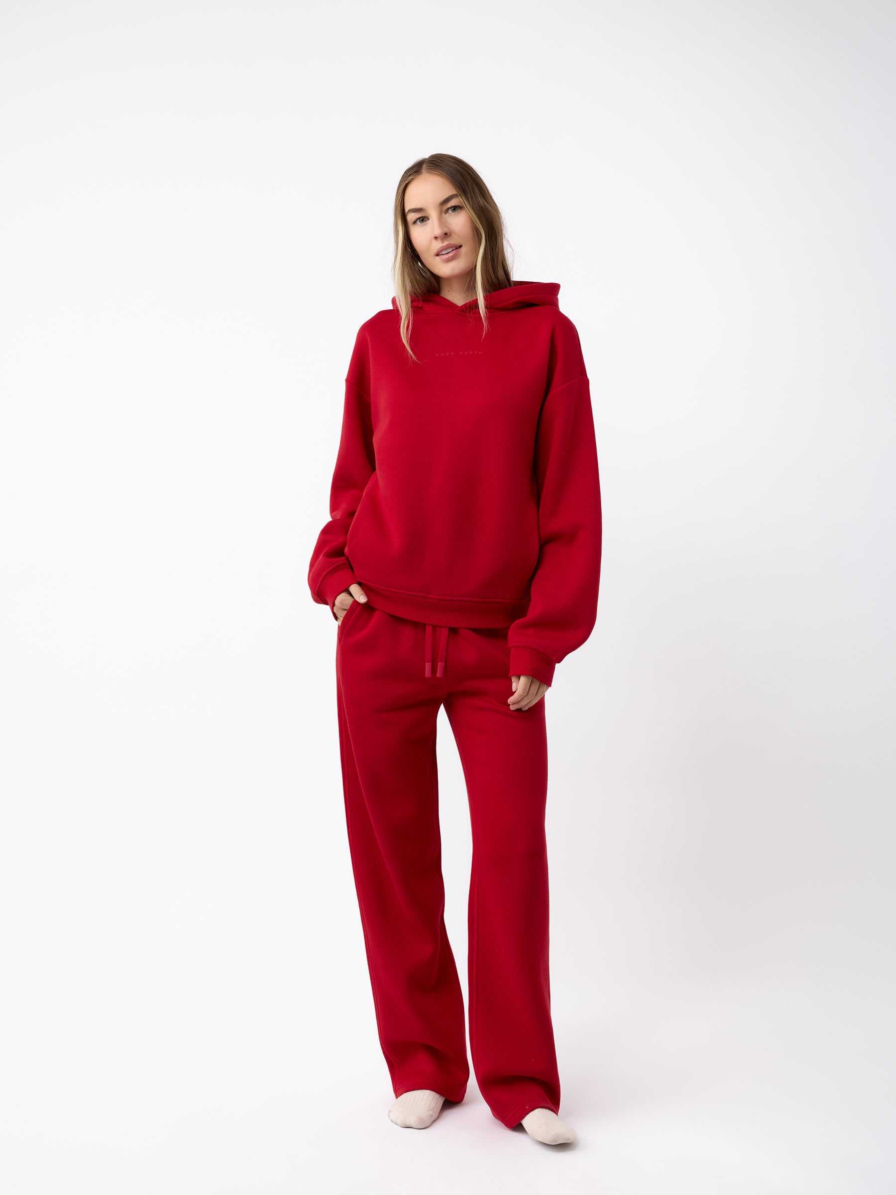 A person stands against a plain white background wearing a matching Women's CityScape Hoodie by Cozy Earth and sweatpants. They have long hair and are looking towards the camera with a relaxed posture. |Color:Crimson
