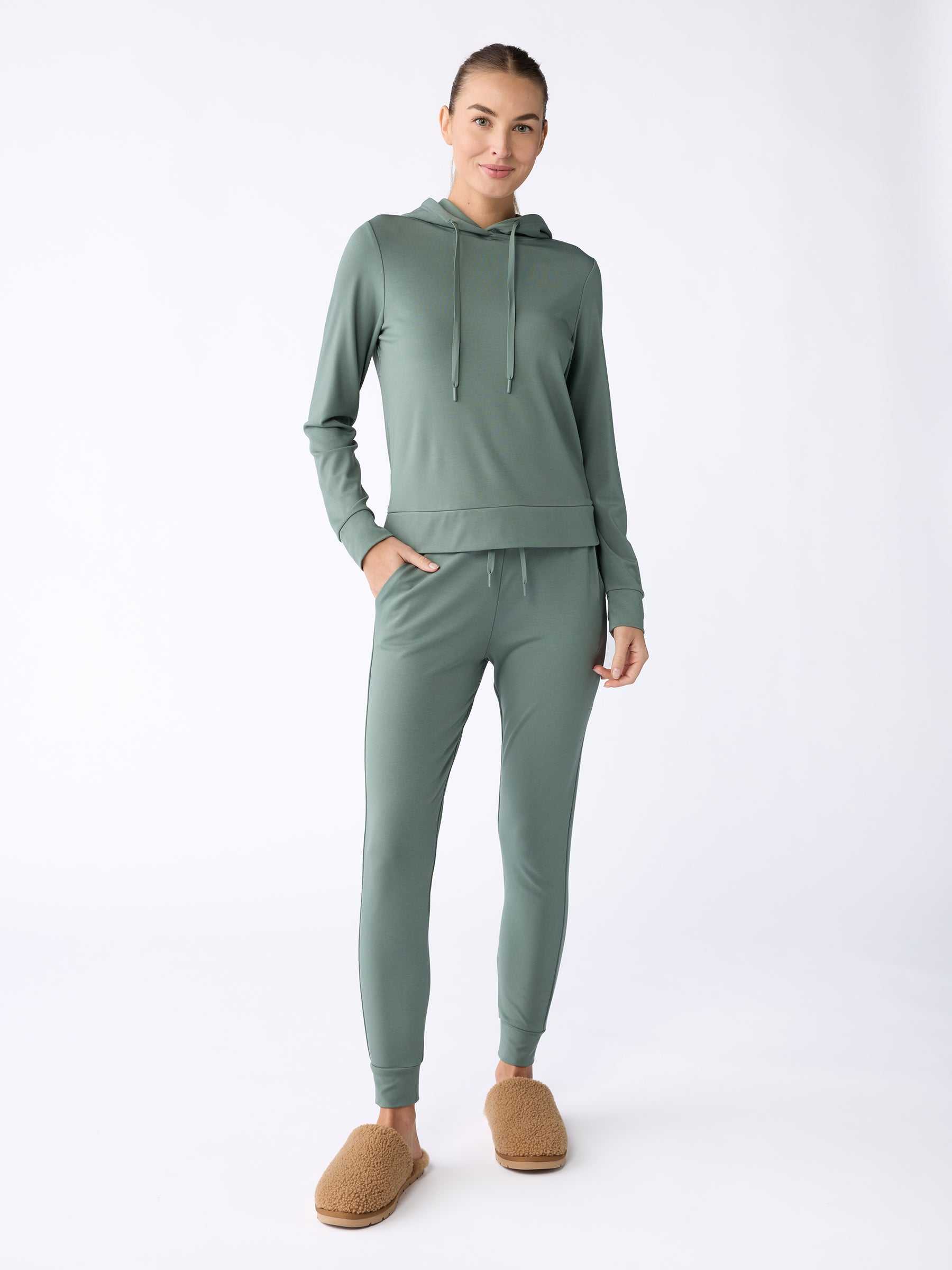 A person stands against a light background, dressed in a matching sage green set from Cozy Earth's Women's Ultra-Soft Bamboo Hoodie collection, with tan slippers. Their hair is styled in a neat bun, hands tucked into pockets, and they are smiling. |Color:Oakmoss