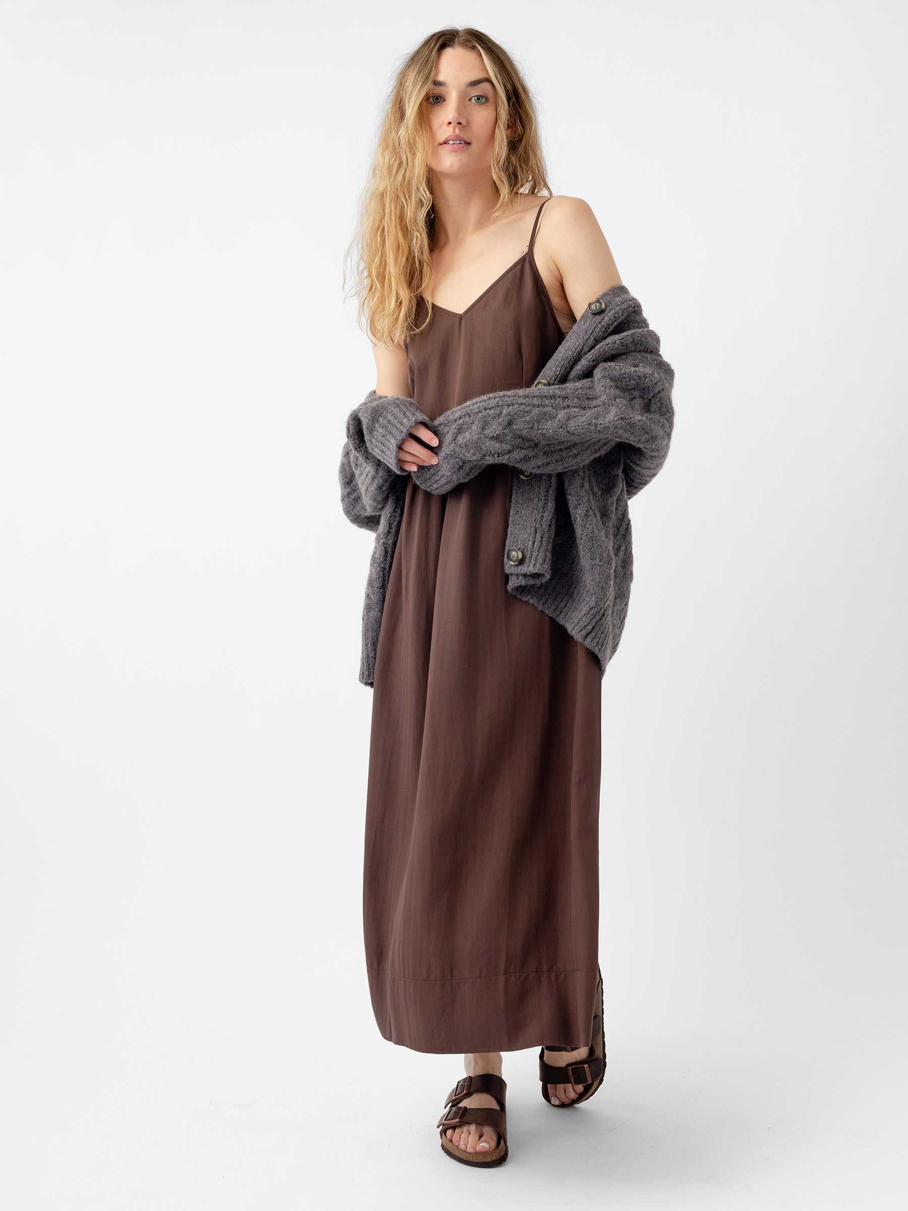 A person with long blond hair is wearing a brown sleeveless dress paired with the Oversized Cable Knit Cardigan from Cozy Earth and sandals, standing against a plain white background. |Color:Thunder