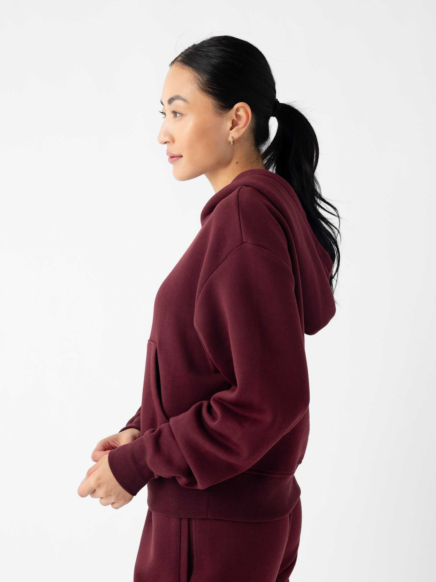 A person with long dark hair tied in a ponytail is seen in a profile view, wearing the Cozy Earth Women's CityScape Cropped Hoodie in maroon and matching pants. The background is plain white, and the person is looking to the left with a relaxed expression. |Color:Burgundy