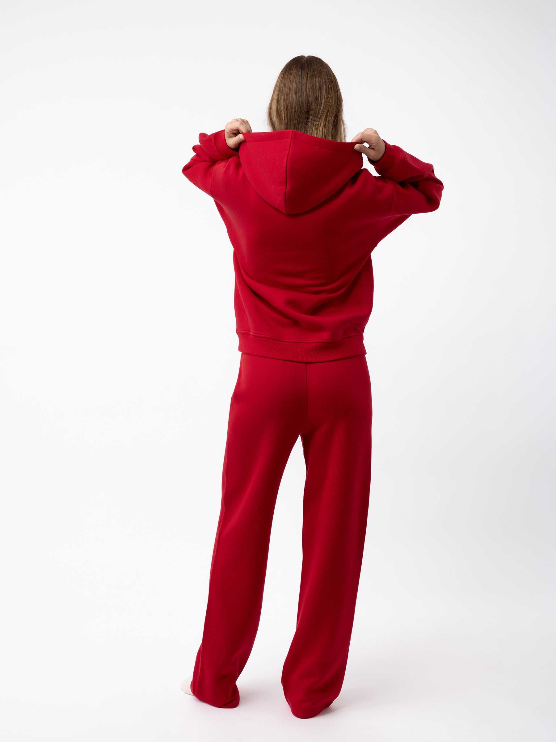 A person stands facing away, wearing a matching red Women's CityScape Hoodie and pants from Cozy Earth. They are holding the hood of the hoodie, set against a plain white background. |Color:Crimson