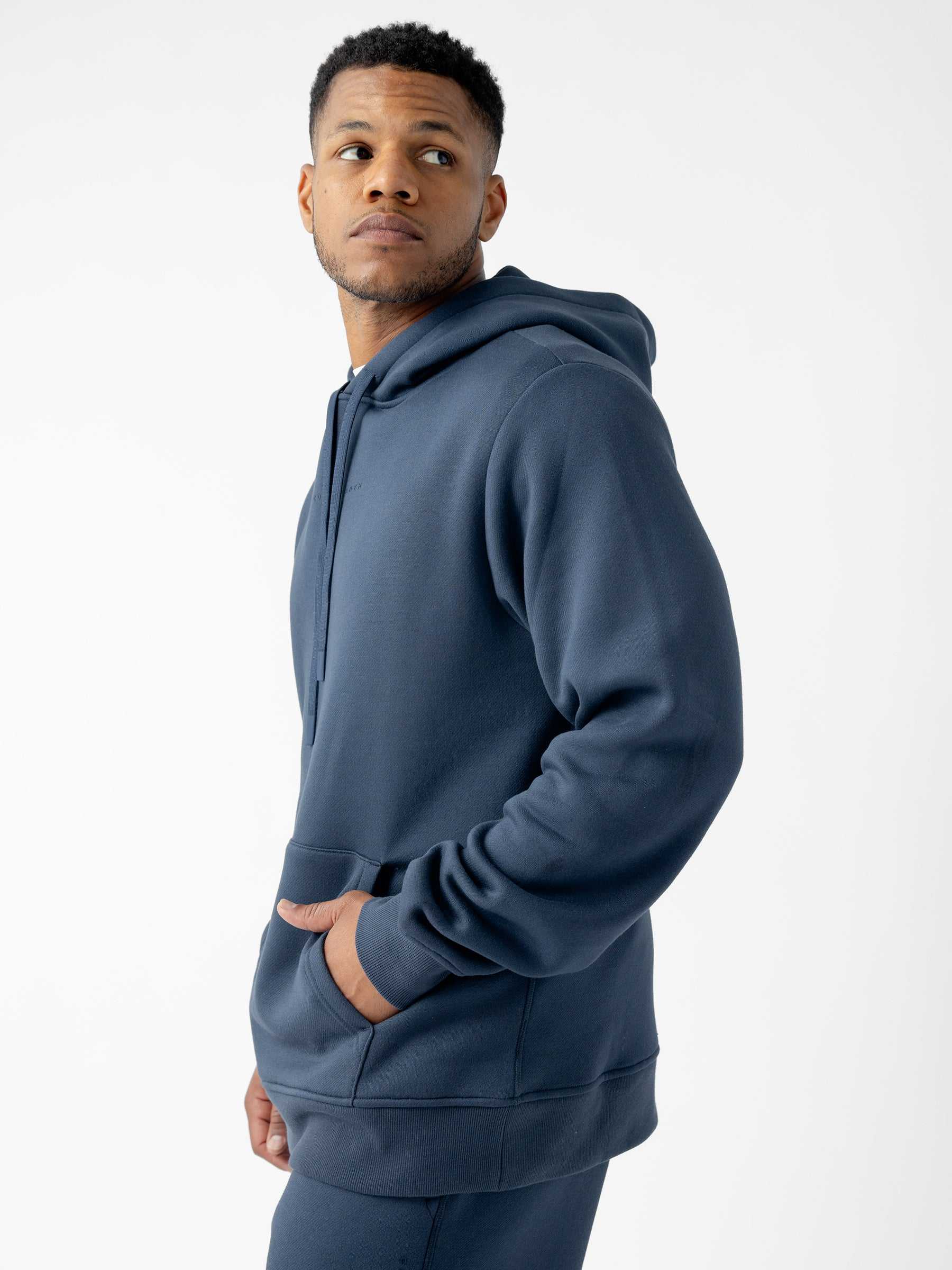 A man wearing a Cozy Earth Men's CityScape Hoodie and sweatpants stands against a plain white background. He is looking off to the side while his left hand is tucked into the hoodie pocket. The dark blue hoodie has a relaxed fit and a drawstring hood. |Color:Eclipse