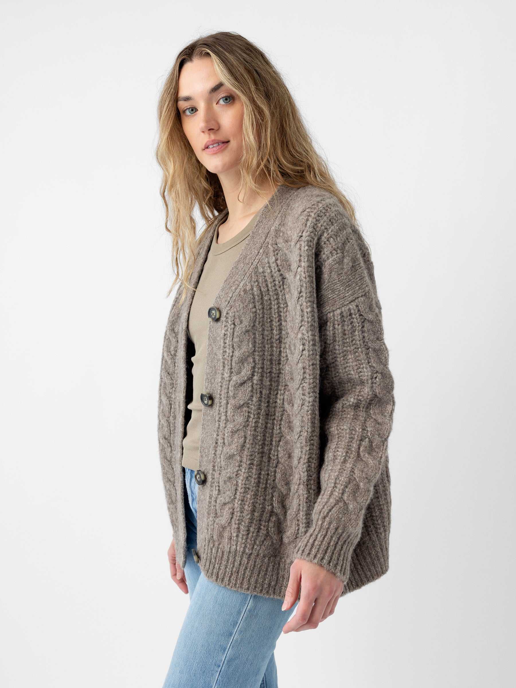 A person with long hair is wearing the "Oversized Cable Knit Cardigan" by Cozy Earth, along with a light shirt. They are standing against a plain background, also dressed in light blue jeans, and gazing toward the camera. |Color:Greige