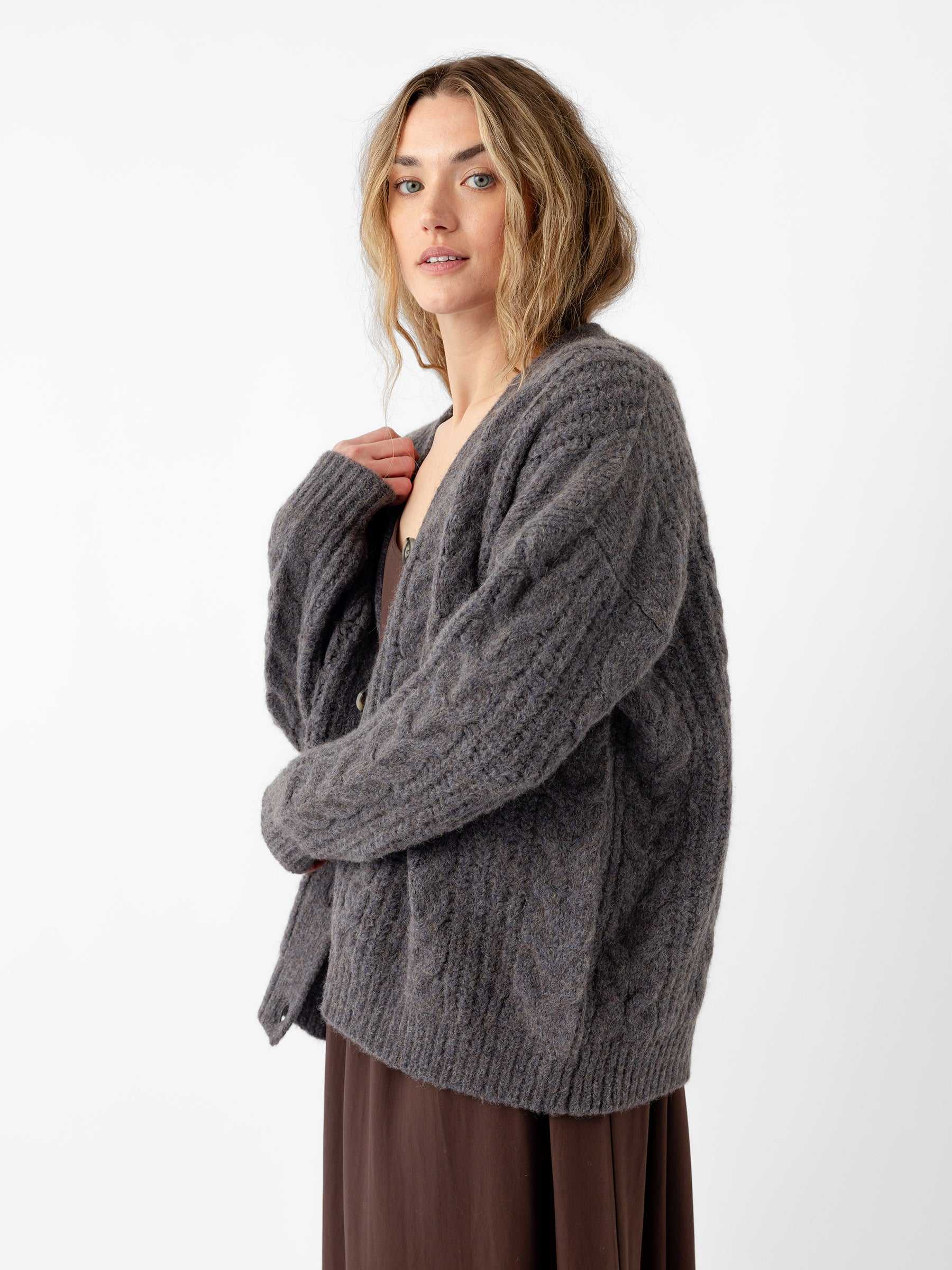 A person with long hair stands against a white background, wearing Cozy Earth's Oversized Cable Knit Cardigan in gray over a brown dress. They maintain a neutral expression as they look towards the camera, gently holding the cardigan collar with one hand. |Color:Thunder