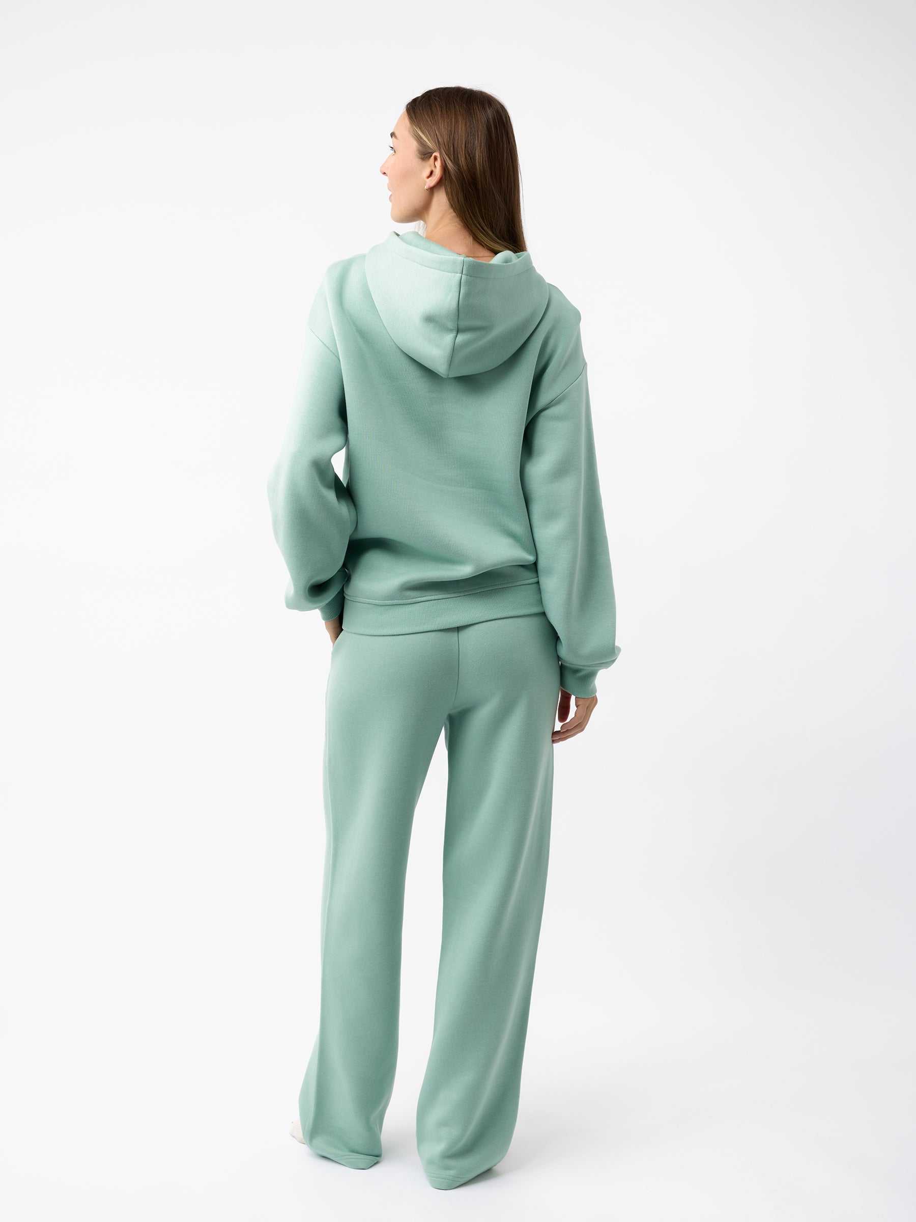 A person with long hair is wearing a green Women's CityScape Hoodie by Cozy Earth, along with matching pants. They are standing against a plain white background, facing away from the camera. |Color:Wintergreen