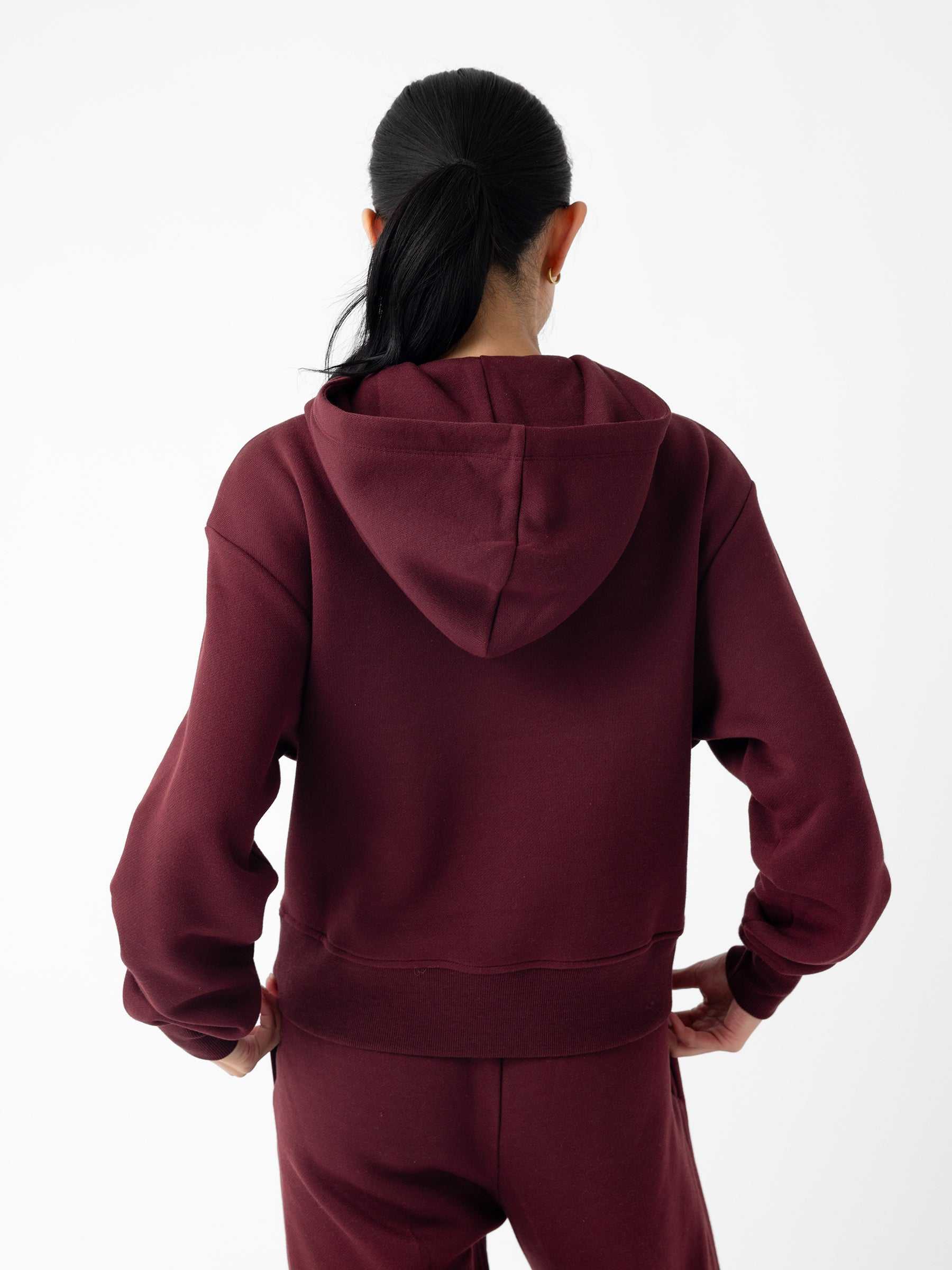 A person with long dark hair in a ponytail, wearing Cozy Earth's Women's CityScape Cropped Hoodie in burgundy and matching pants, stands with hands on hips. The person is facing away, showcasing the back of the outfit against a plain white background. |Color:Burgundy