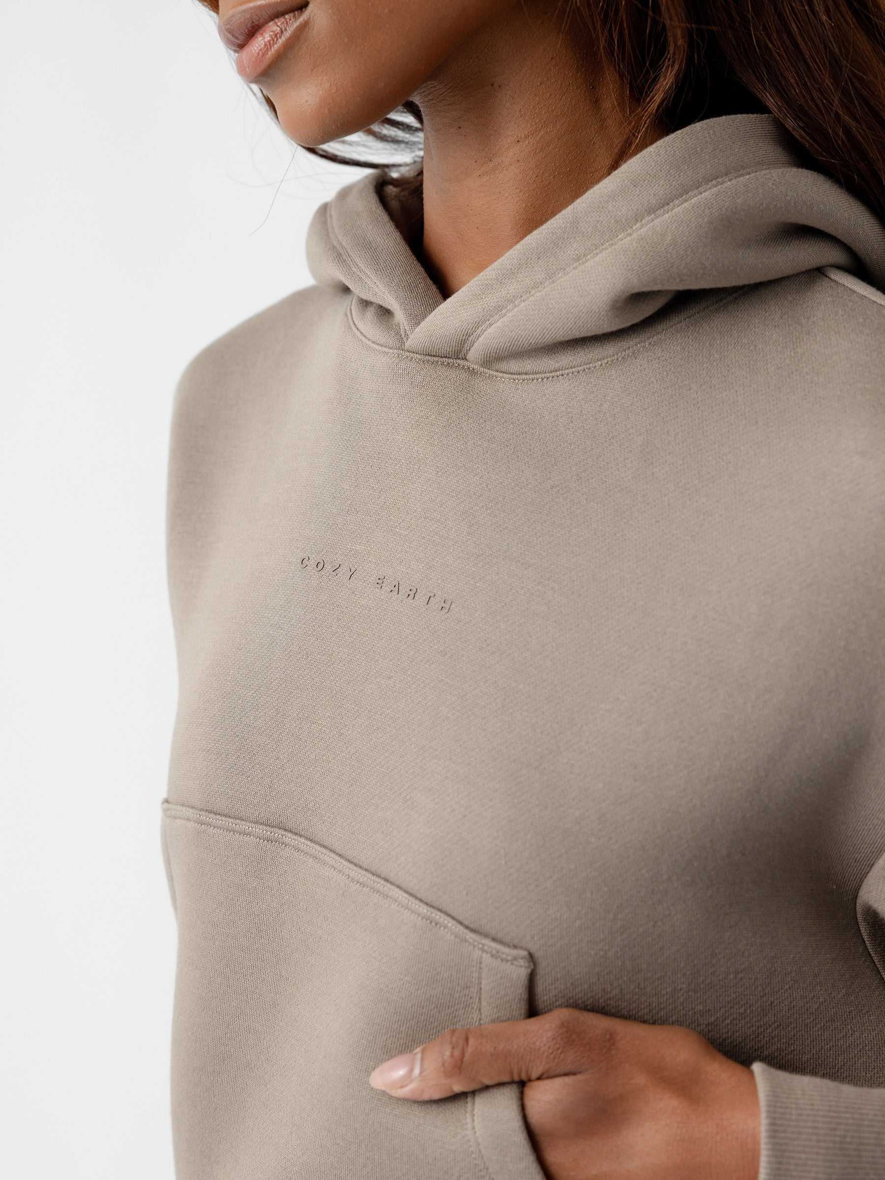 A close-up shot captures a person wearing the Women's CityScape Cropped Hoodie by Cozy Earth. The lower part of their face, neck, and upper torso are shown. This beige hoodie features a front pocket and text embroidered on the chest. The individual’s hand rests inside the pocket, against a plain white background. |Color:Ebony