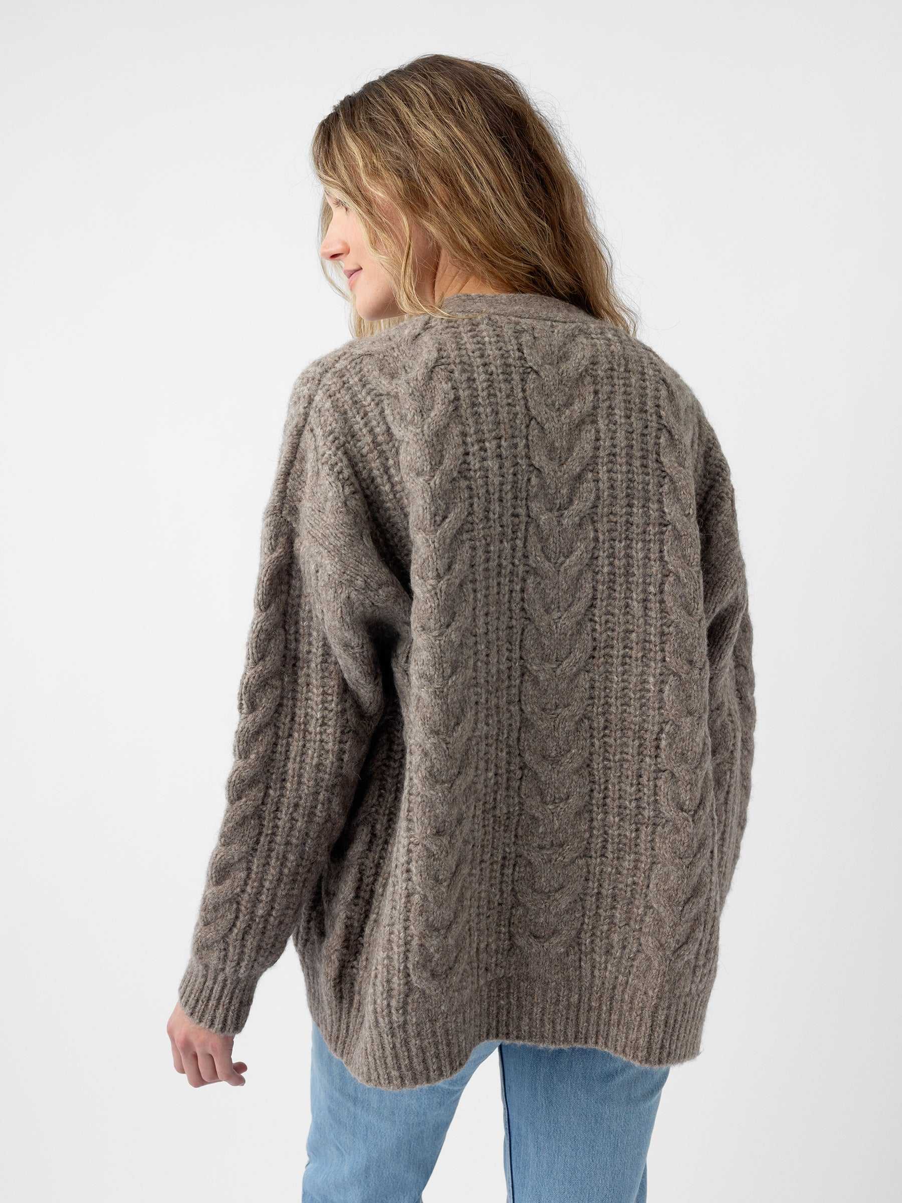 A person with long hair is wearing a grey Oversized Cable Knit Cardigan from Cozy Earth and blue jeans, standing against a plain white background with their head slightly turned to the side. |Color:Greige