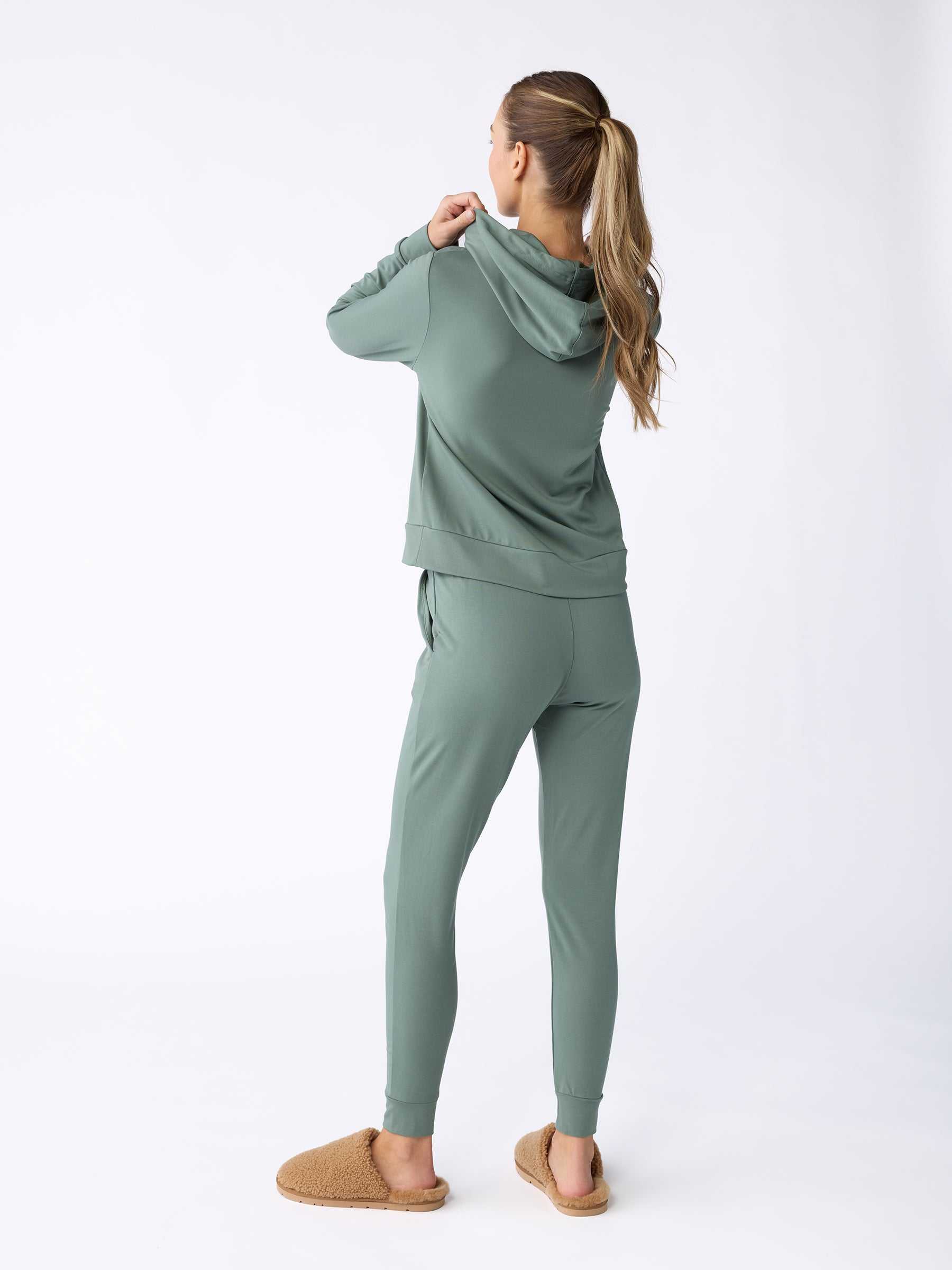 A woman with long hair stands facing away from the camera on a white background, wearing a cozy green Women's Ultra-Soft Bamboo Hoodie from Cozy Earth and tan slippers. |Color:Oakmoss