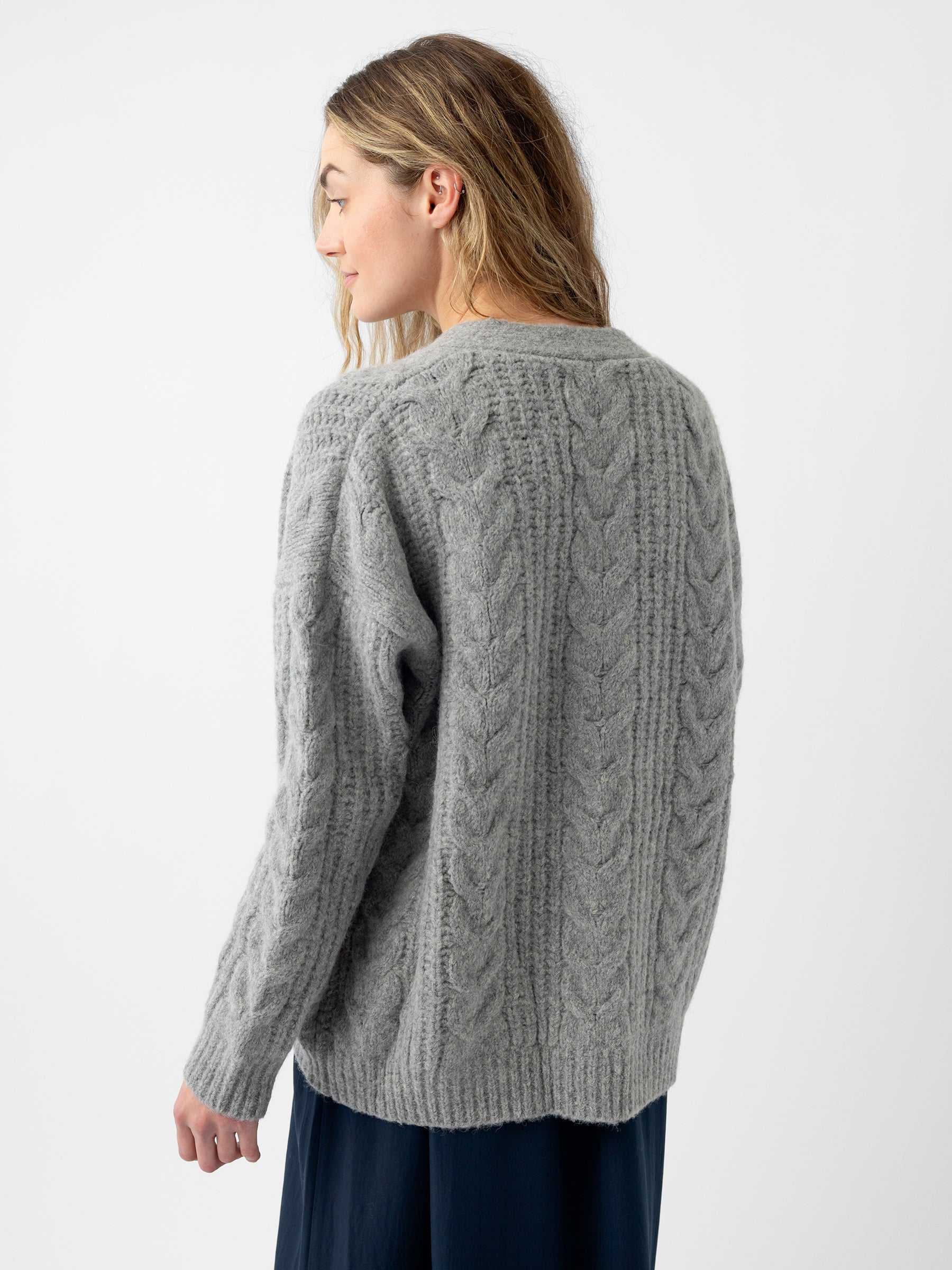 A woman with long hair is wearing a gray Oversized Cable Knit Cardigan from Cozy Earth. She is facing away, with her head turned slightly to the side, against a plain white background. The bottom of her skirt peeks into view at the edge of the image. |Color:Oxford
