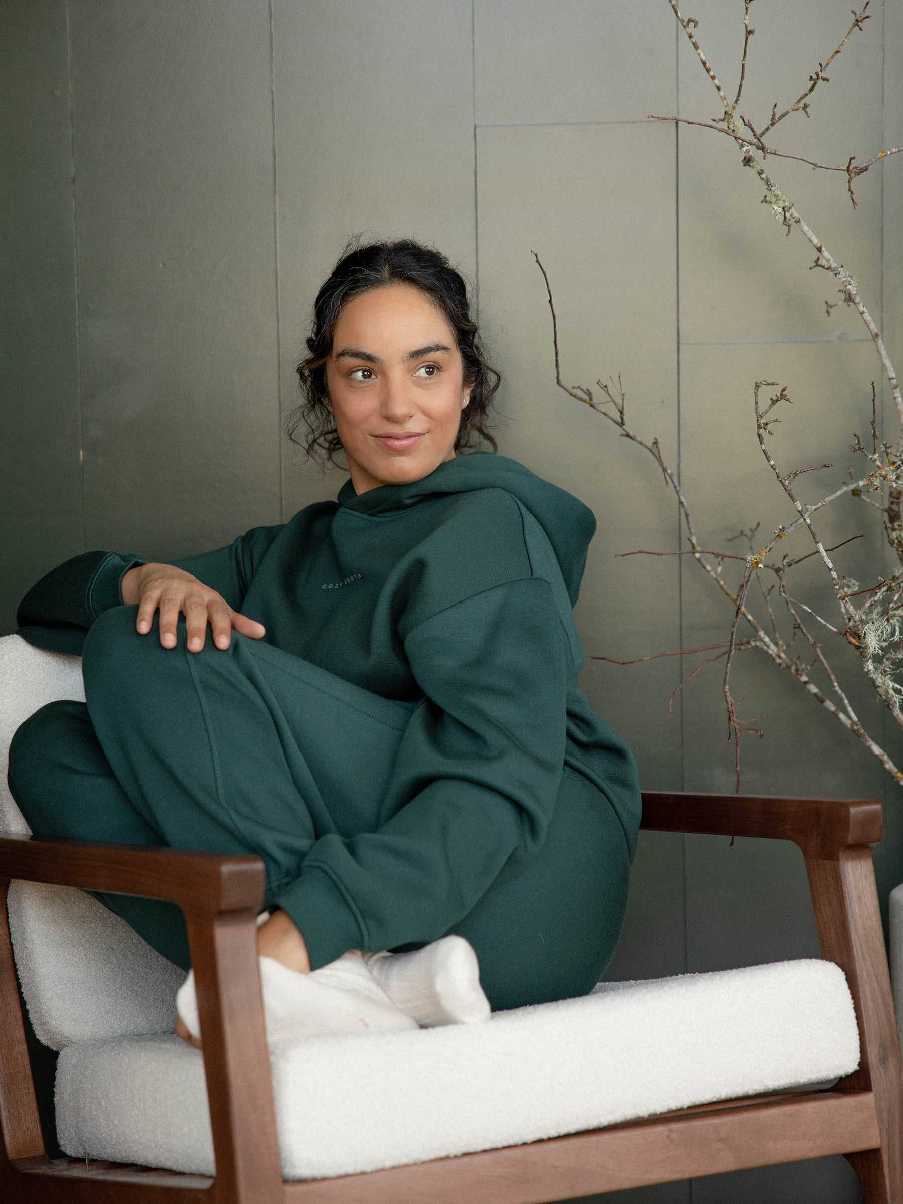 A person with dark hair sits comfortably on a white cushioned chair, wearing the Cozy Earth's Women's CityScape Hoodie paired with sweatpants. They have a relaxed expression as they gaze off-camera. In the background, a bare branch decorates the scene against a greenish wall. |Color:Spruce