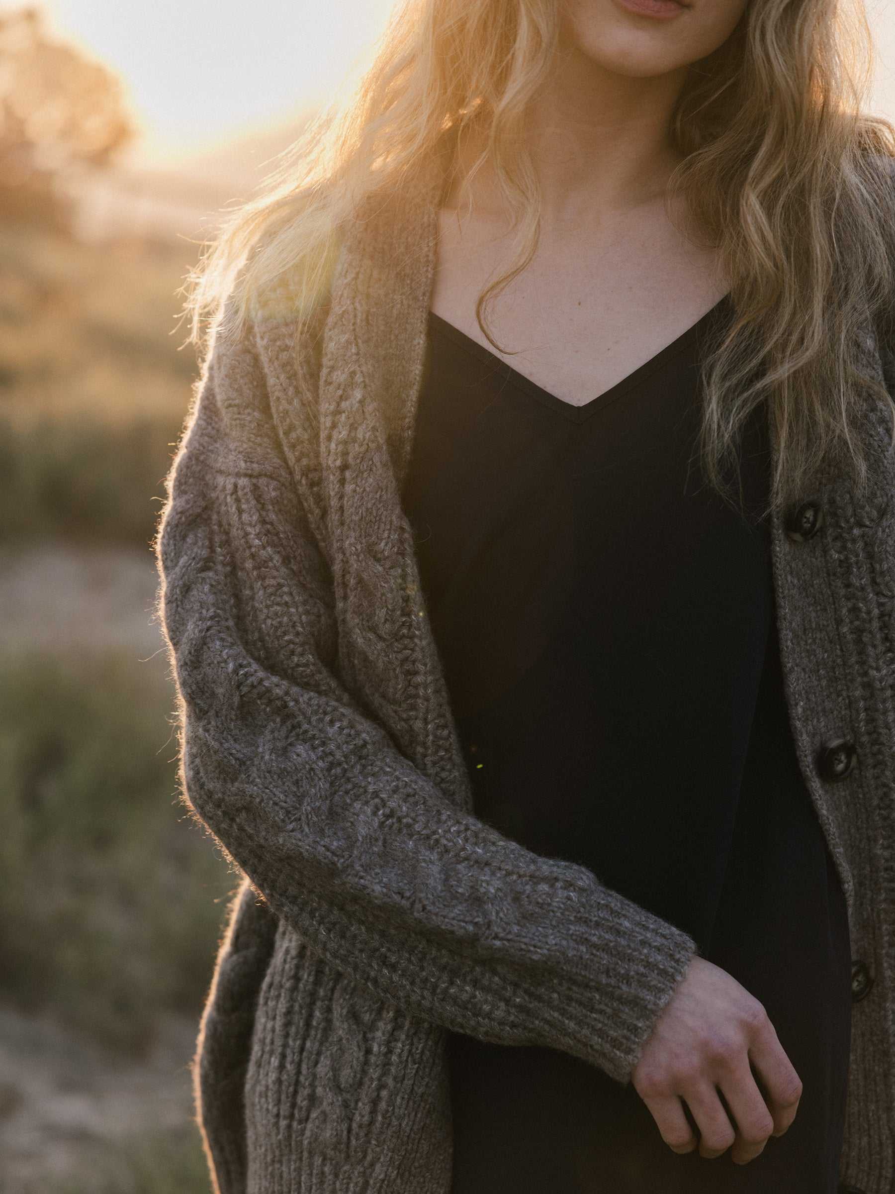 A woman with long blonde hair stands outdoors during golden hour, wearing a black dress and an Oversized Cable Knit Cardigan from Cozy Earth. The warm sunlight creates a soft glow, highlighting the natural setting with grass in the background. |Color:Greige