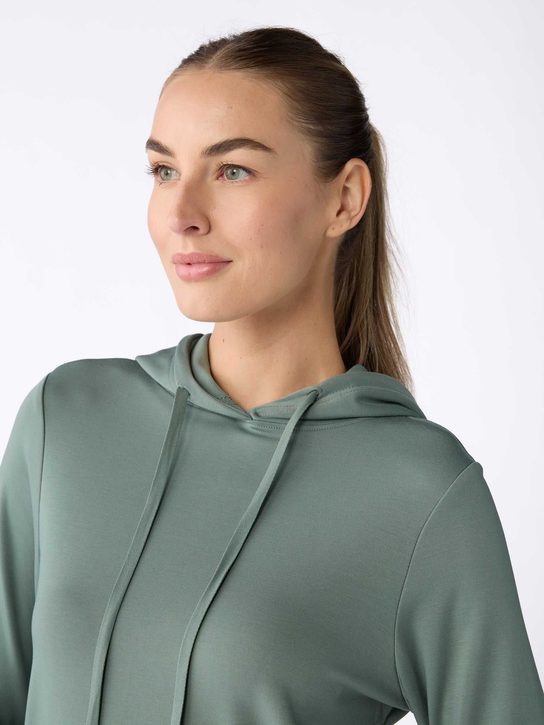 A person with long hair is looking to the side while wearing a Women's Ultra-Soft Bamboo Hoodie in sage green from Cozy Earth. The background is plain white. |Color:Oakmoss