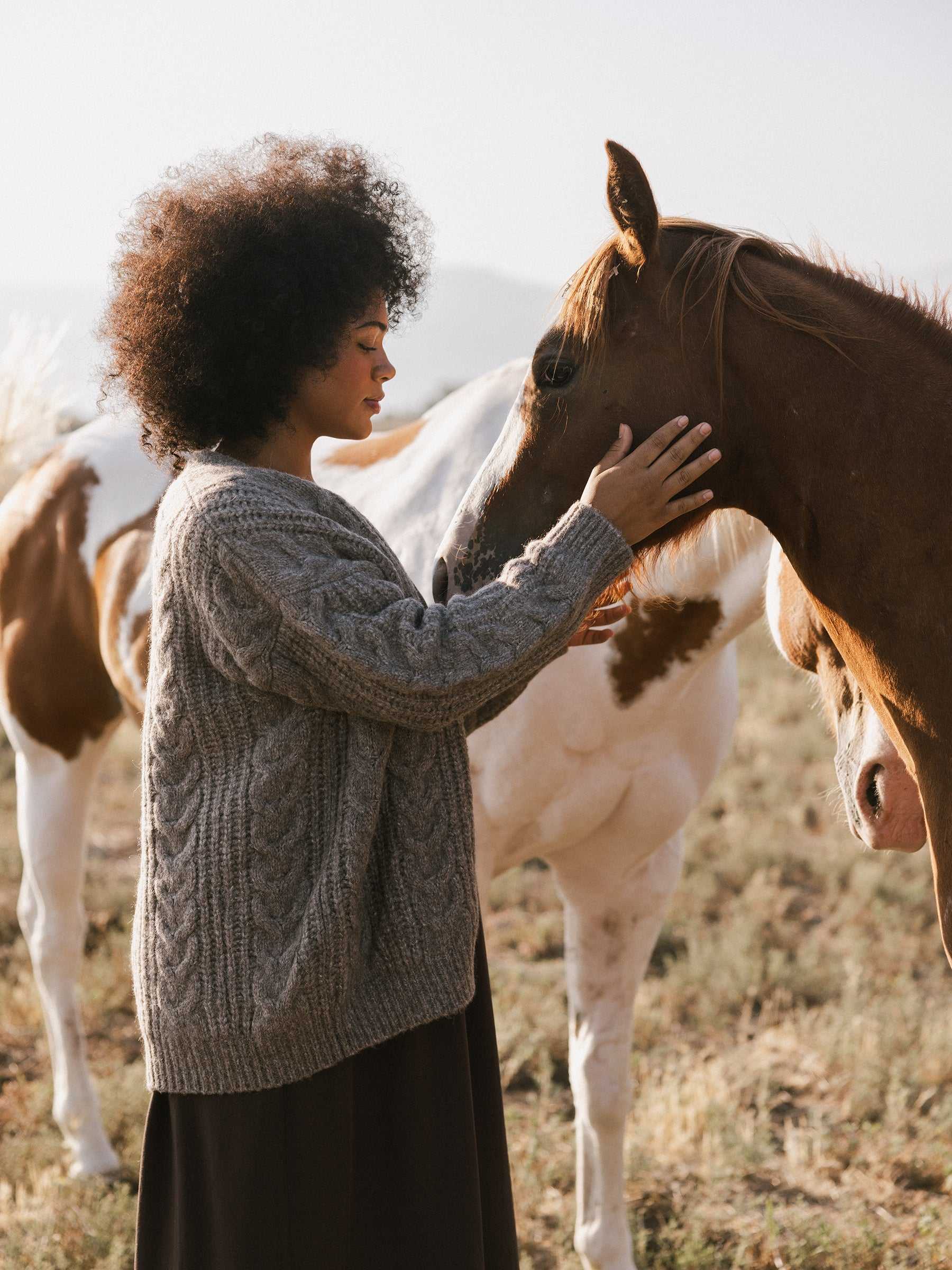 A person stands in a field, softly touching the nose of a horse with a white mane. They are wearing an Oversized Cable Knit Cardigan by Cozy Earth, and there are other horses in the background. The scene is peaceful and serene. |Color:Oxford