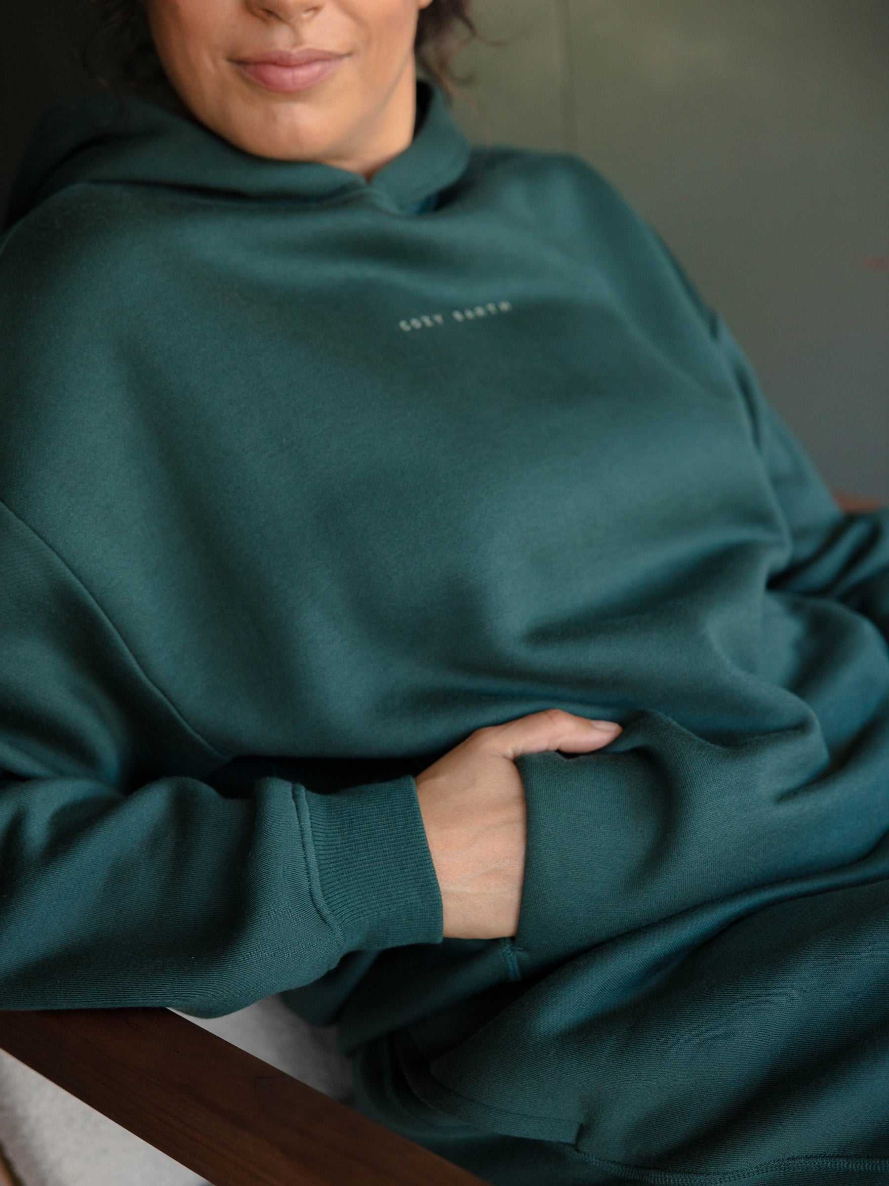 A person wearing a Cozy Earth Women's CityScape Hoodie in teal, featuring subtle chest text, sits in a chair with their left hand tucked into the hoodie's pocket against a softly blurred background. |Color:Spruce