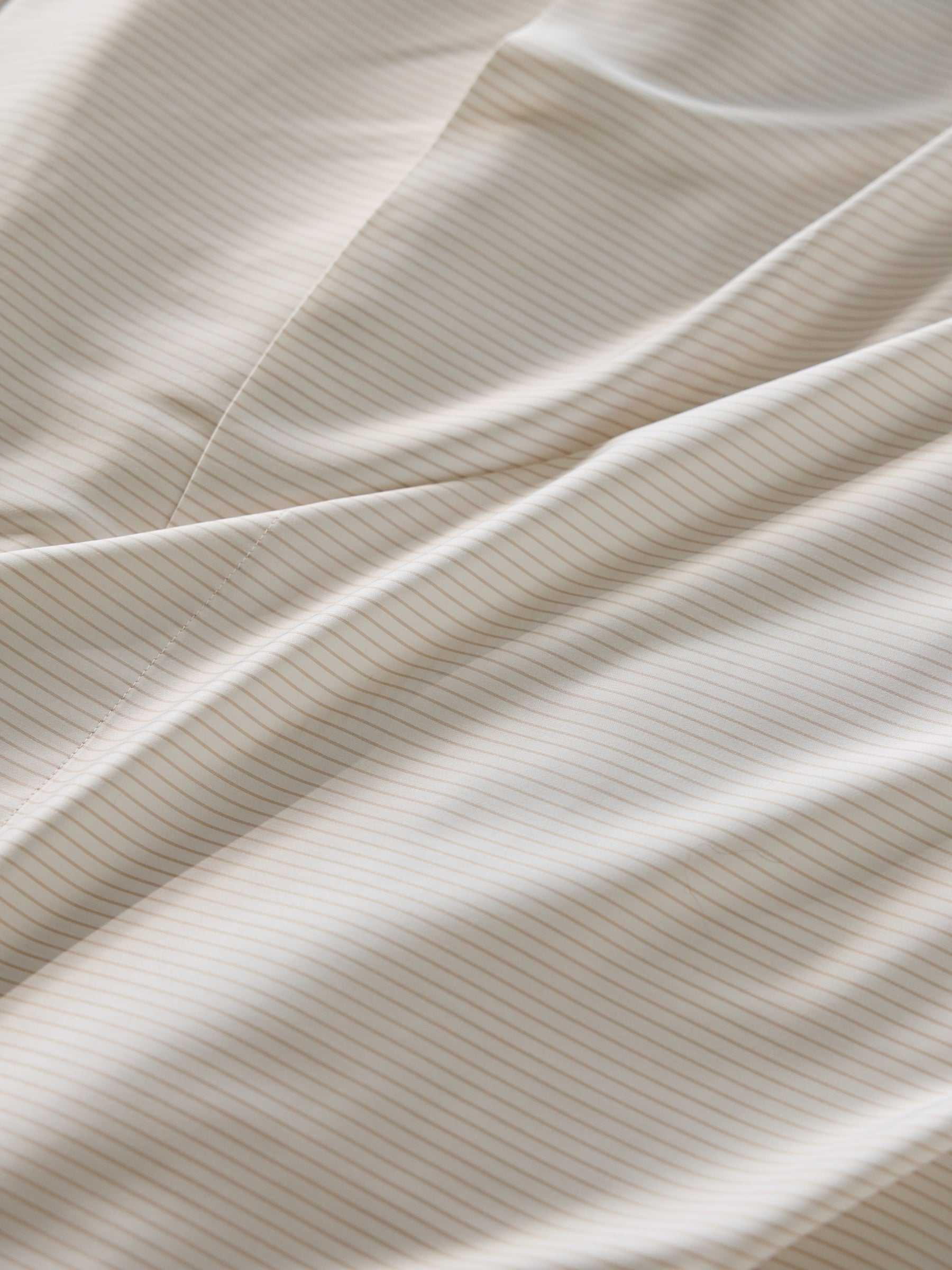 Close-up of the Bamboo Duvet Cover by Cozy Earth, showcasing its smooth, white fabric adorned with thin, light beige stripes running diagonally. The duvet cover is gently folded, creating soft waves and subtle shadows. |Color:Driftwood Pinstripe