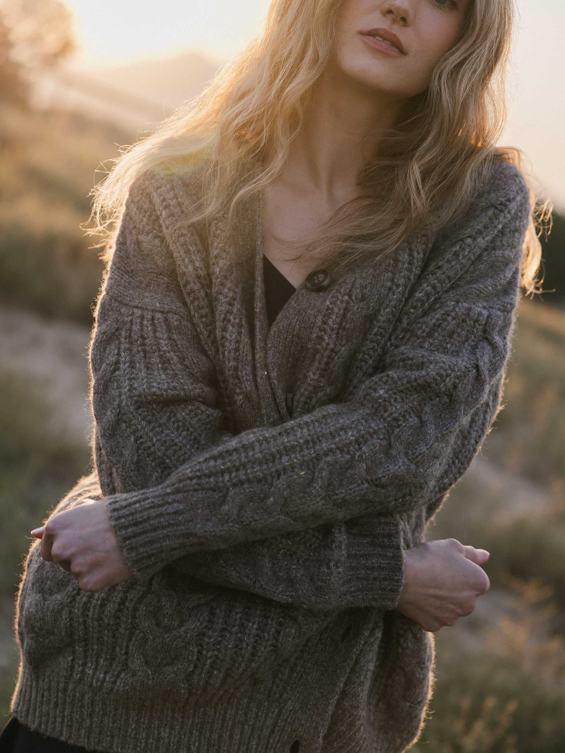 A person with long, wavy hair stands outdoors in warm lighting, wrapped in a Cozy Earth Oversized Cable Knit Cardigan. The background reveals a blurred natural setting, evoking a tranquil, autumnal ambiance. |Color:Greige