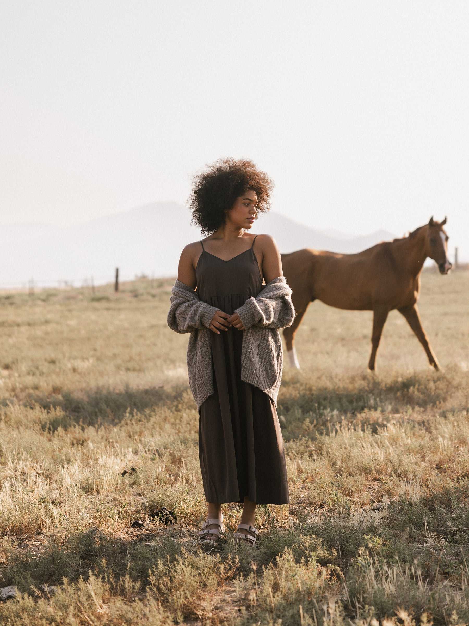 A woman stands in a grassy field wearing a long dark dress and an Oversized Cable Knit Cardigan by Cozy Earth. A horse grazes in the background, with the scene bathed in soft, natural light and mountains visible in the distance. |Color:Oxford