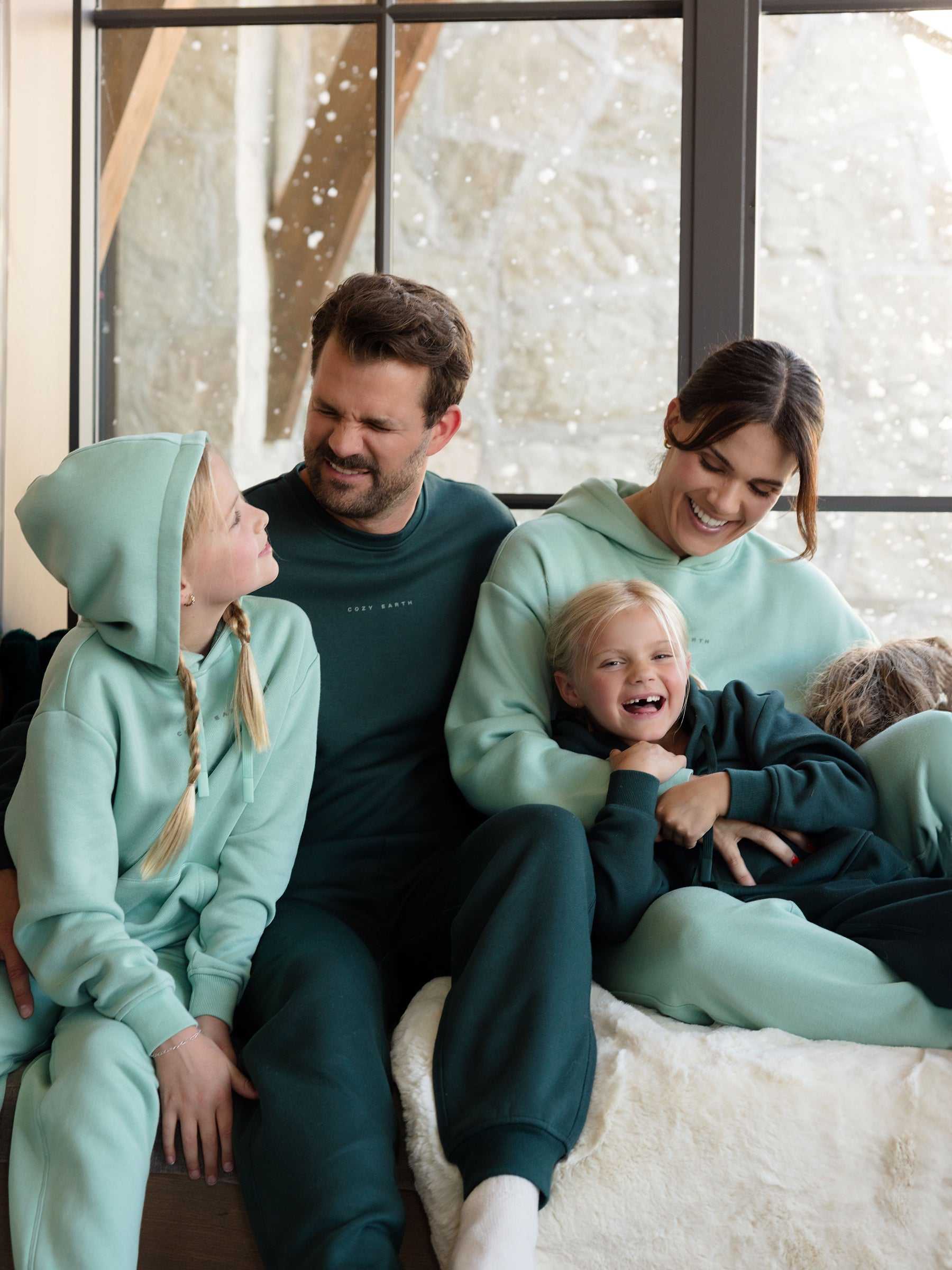 A family of four is gathered indoors, smiling as they wear matching teal and green Women's CityScape Hoodies from Cozy Earth, paired with sweatpants. The cozy and cheerful scene is complete with a snowy landscape visible through the large windows in the background. |Color:Wintergreen
