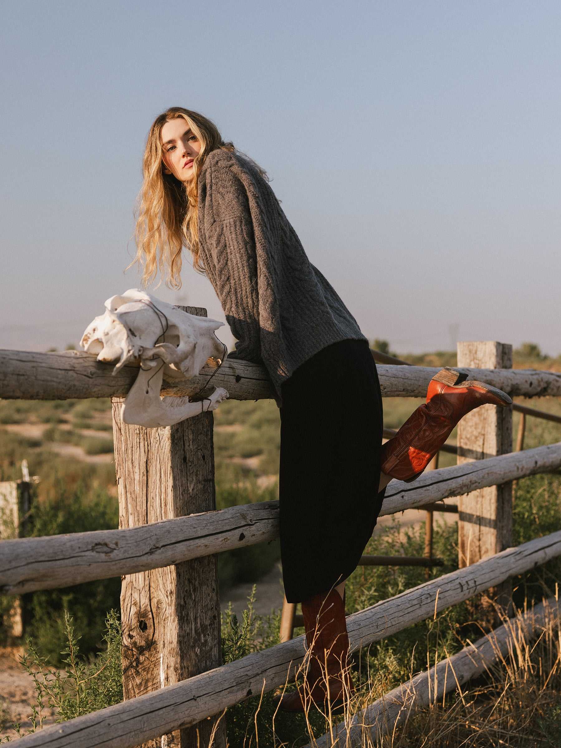 A person with long hair, dressed in an Oversized Cable Knit Cardigan by Cozy Earth, a black skirt, and red boots, leans on a wooden fence while gazing into the distance. A white skull is positioned next to them in this rural setting under a clear blue sky. |Color:Thunder