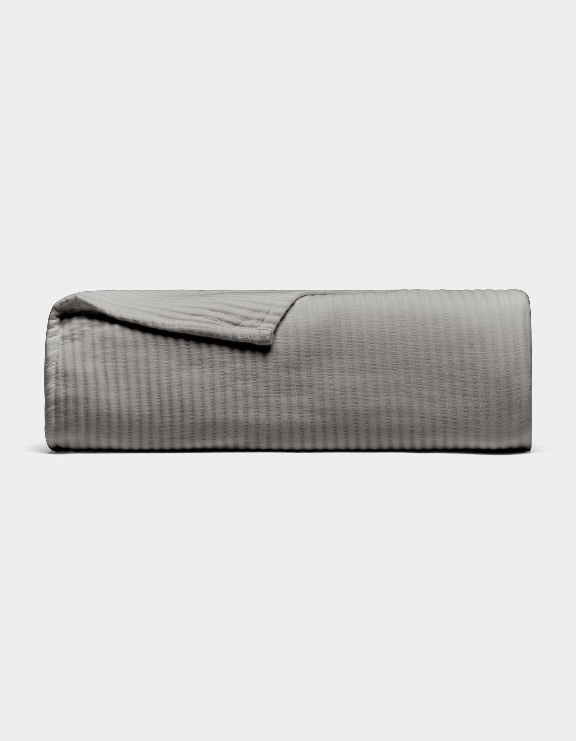 The Cozy Earth Bamboo Coverlet, shown neatly folded in light gray with a subtle textured pattern, is displayed on a plain white background. One corner is turned down to reveal the inner side of the coverlet.|Color:Charcoal