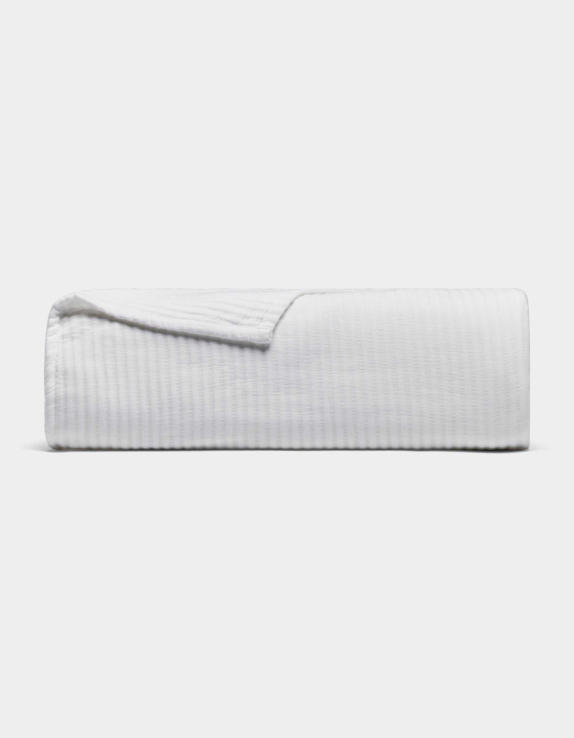 The Cozy Earth Bamboo Coverlet, a white textured blanket, is neatly folded with one corner slightly lifted, set against a plain light gray background.|Color:White