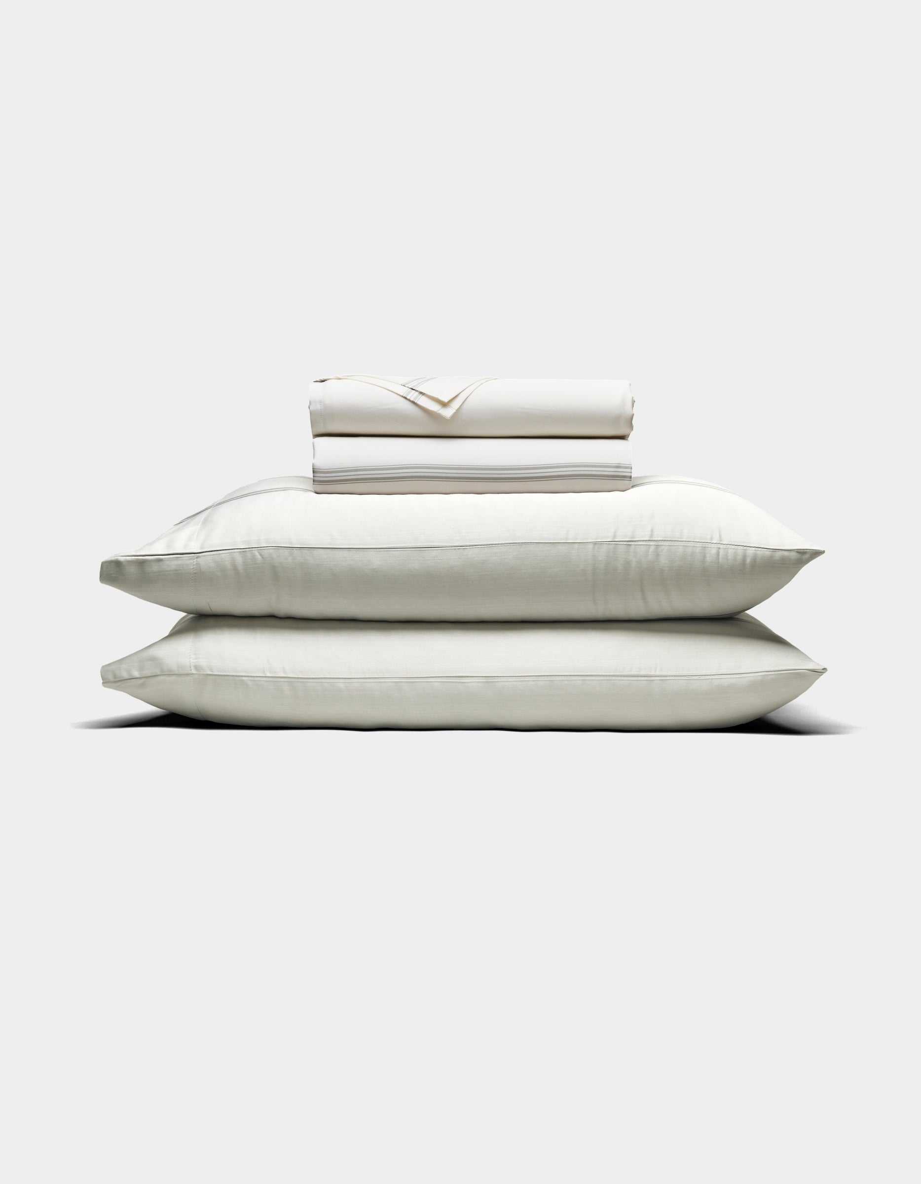 Two large white pillows are stacked with a folded Cozy Earth Bamboo Sheet Set and pillowcases placed on top, all arranged against a plain light background.|Color:Baja Stripe in Laurel