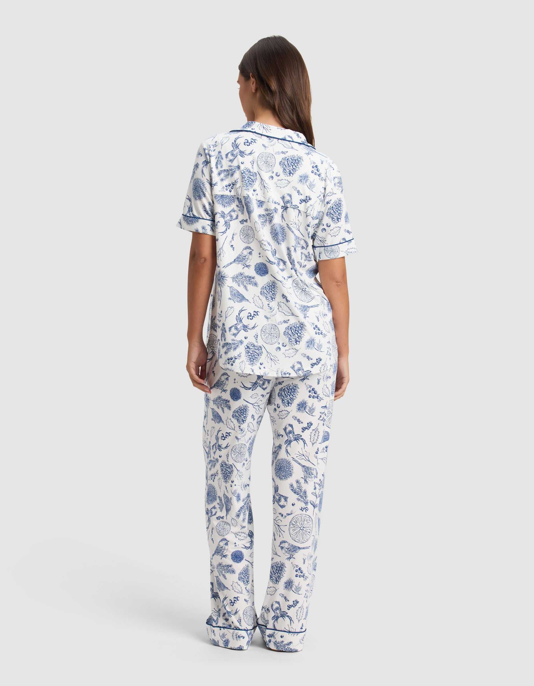 A woman with long brown hair faces away, wearing the Cozy Earth Women's Bamboo Stretch-Knit Short Sleeve Pajama Set in a blue and white botanical pattern with navy trim, set against a plain light gray background.|Color:Winter Toile Pacific