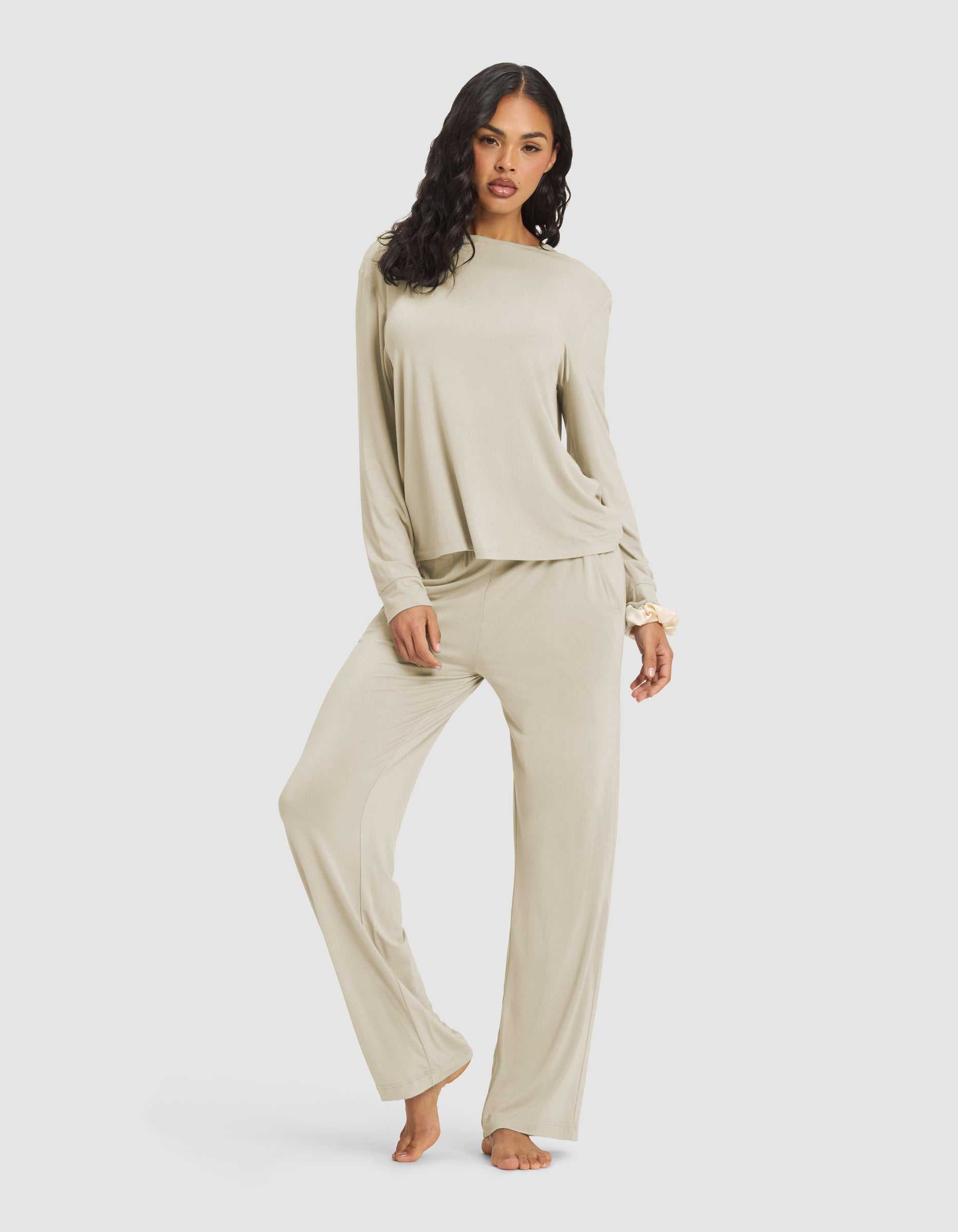 A woman with long, wavy dark hair models the Cozy Earth Women's Loews Hotel Lounge Set—a loose, light beige pajama set with long sleeves and wide-leg pants—as she stands barefoot against a plain white background.