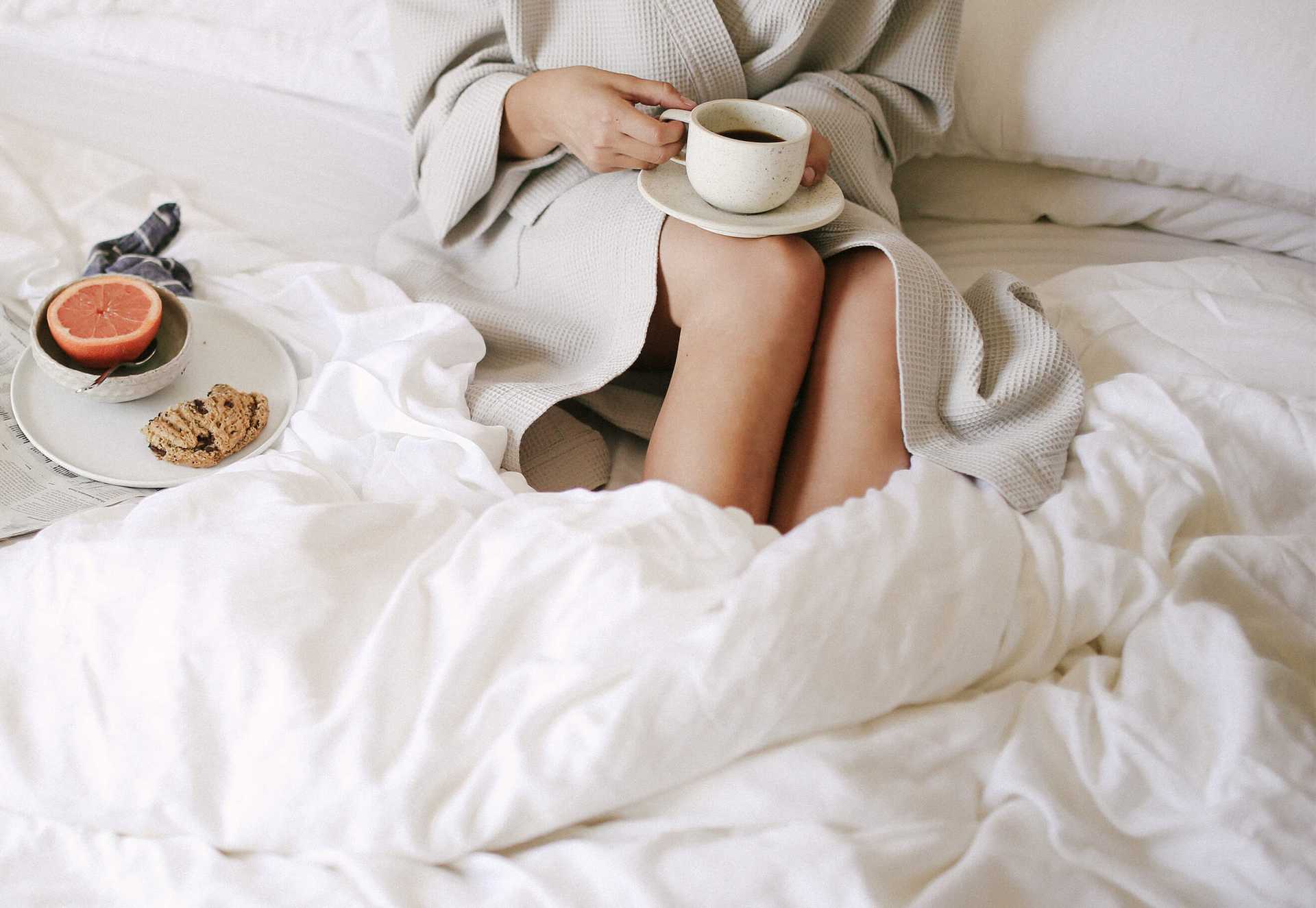 Person in waffle robe sitting on bed with white sheets and a cup of coffee |Color:White