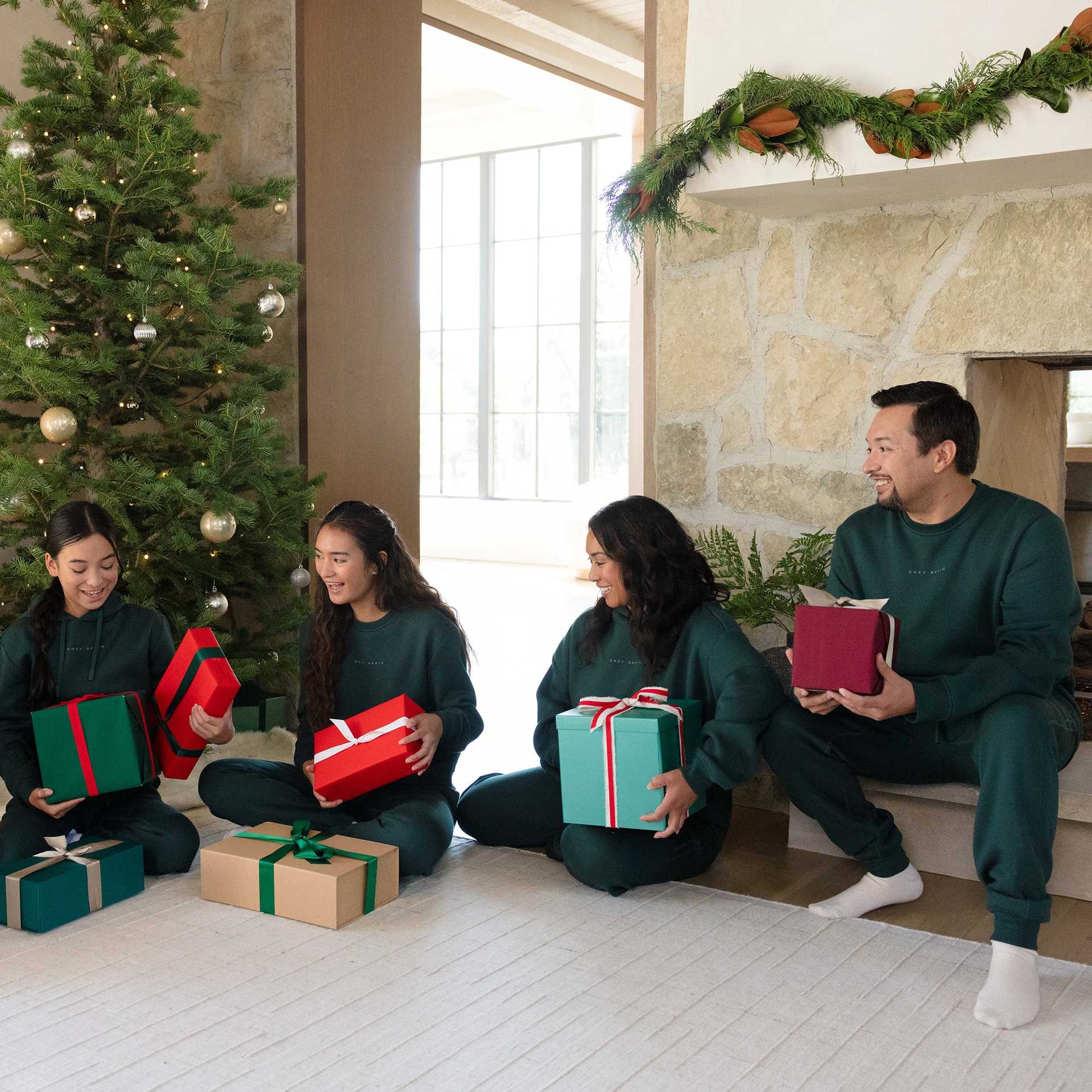A family of four, dressed in matching green Men's CityScape Crewnecks by Cozy Earth, sits on the floor near a beautifully decorated Christmas tree and stone fireplace. They are smiling and holding wrapped gifts in various colors, while garland elegantly decorates the mantel. |Color:Spruce