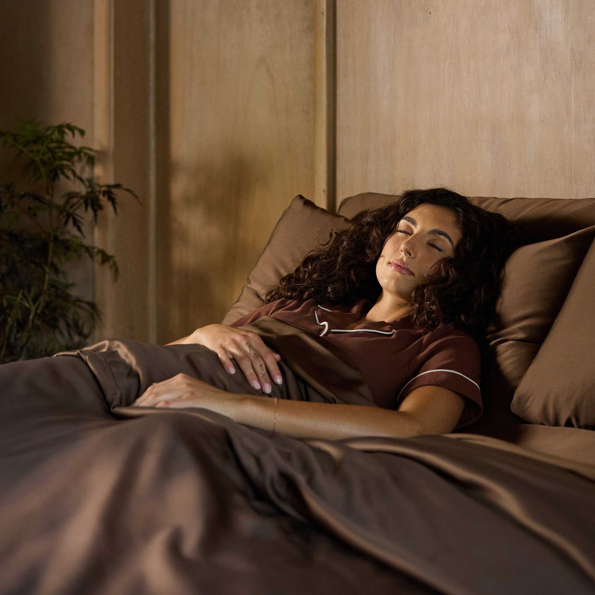 A person with long, curly hair is lying in bed asleep, wearing brown pajamas. They are covered with a Cozy Earth Bamboo Duvet Cover, and a wooden wall panel background is visible. A plant is seen on the left side of the image. |Color:Coffee