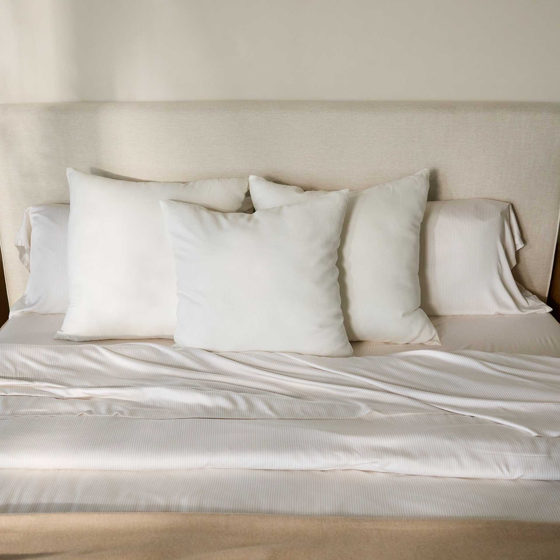A neatly made bed, featuring a beige fabric headboard, is adorned with four white pillows and topped with the Bamboo Duvet Cover by Cozy Earth. Sunlight softly illuminates the bedding, creating a serene and inviting atmosphere. |Color:Driftwood Pinstripe