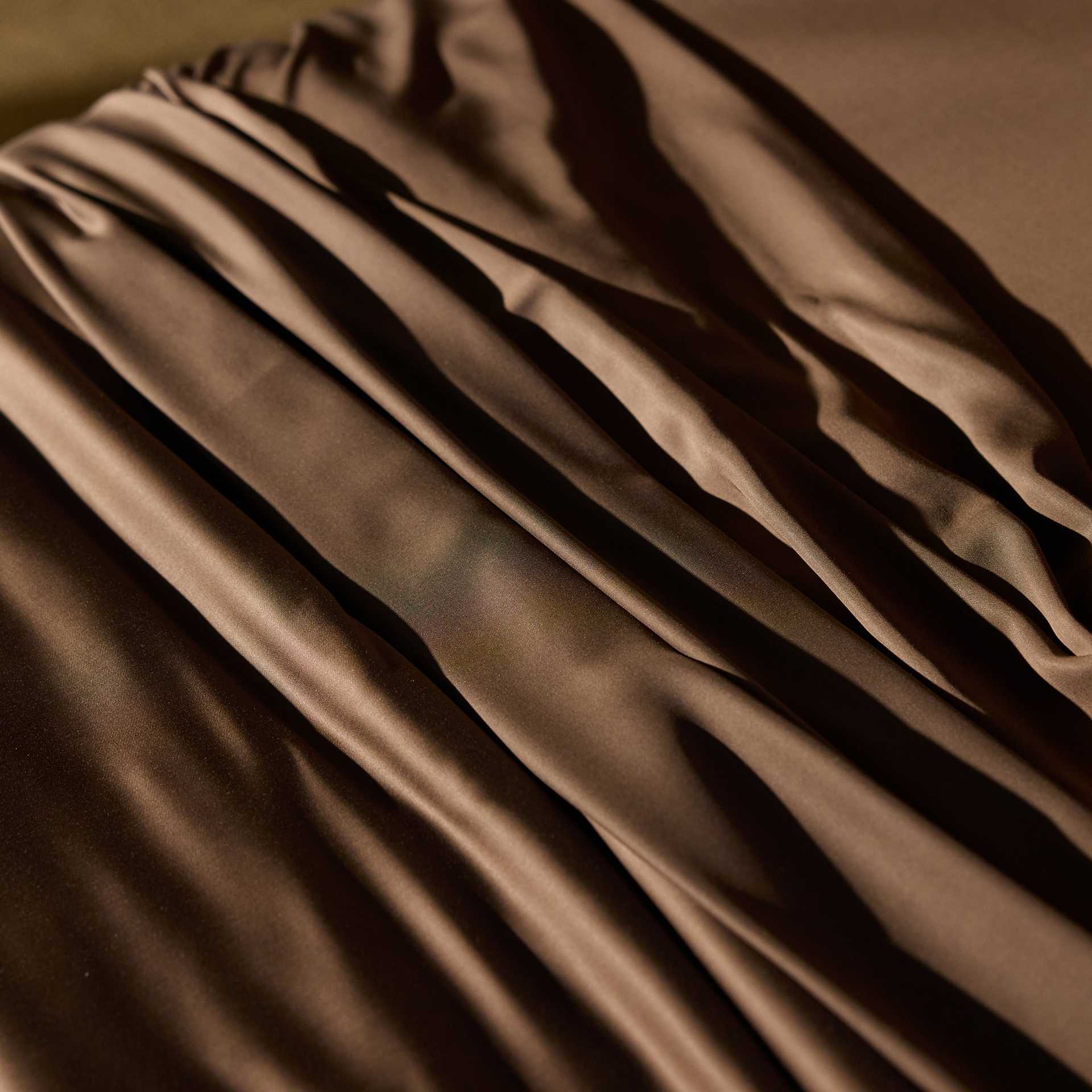 A close-up of the Bamboo Duvet Cover by Cozy Earth, showcasing its smooth, brown satin fabric draped in gentle folds. The texture of the material reflects light, creating a soft sheen and highlighting the fluid lines of the cover's arrangement. |Color:Coffee