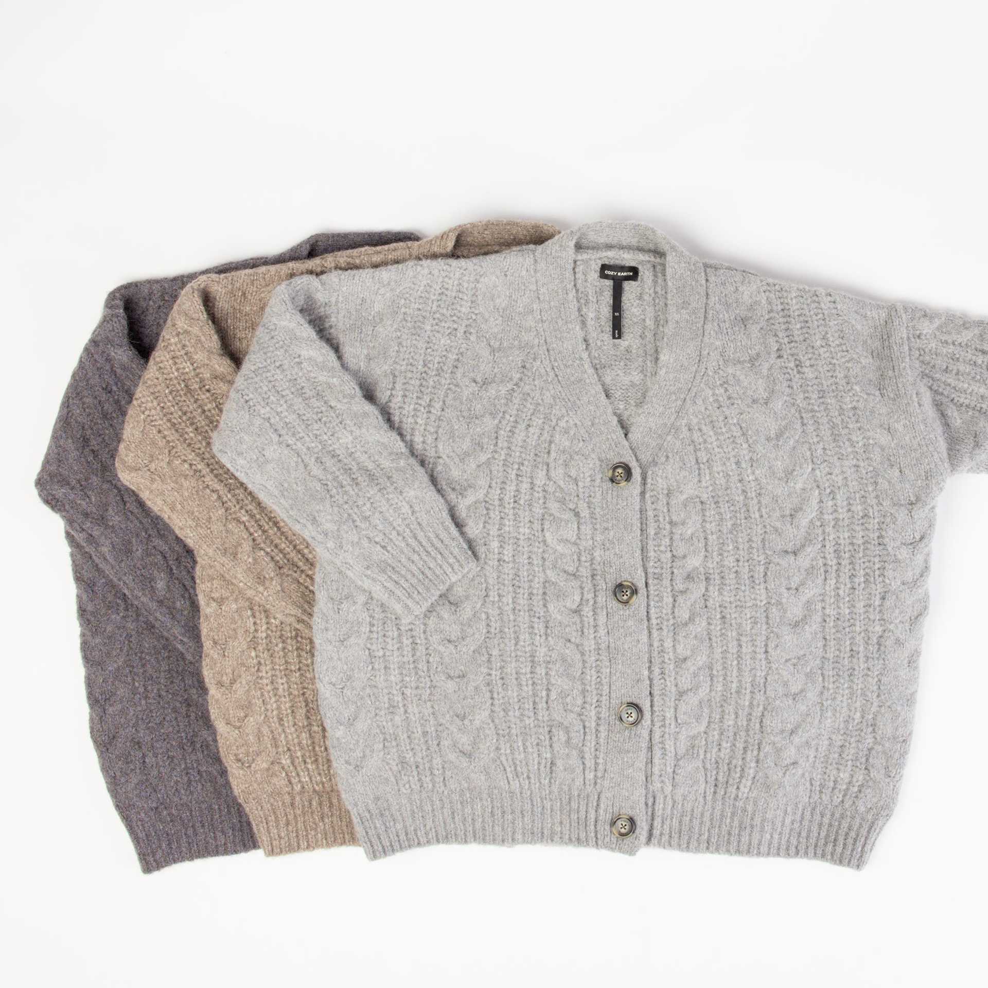 Three Oversized Cable Knit Cardigans by Cozy Earth, in gray, beige, and dark gray, are neatly folded and stacked on a white background. Each cardigan features a V-neck design and button closure. |Color:Greige
