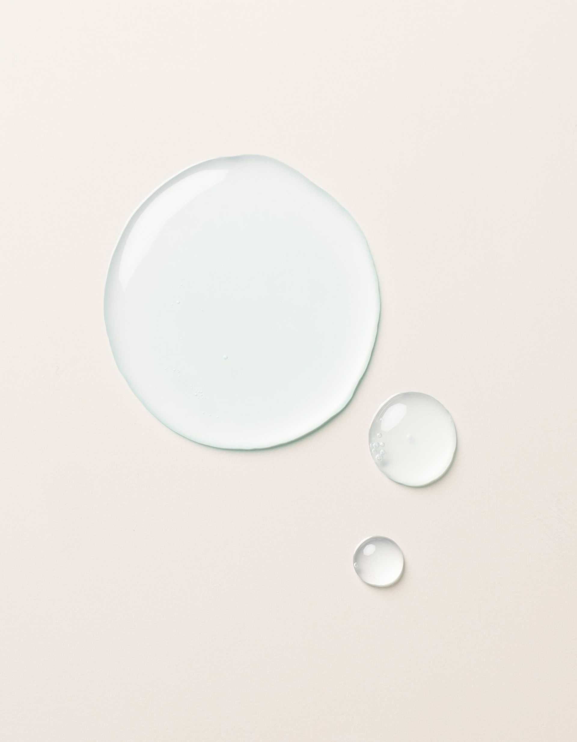 The Hydration Duo by Cozy Earth is shown as a large drop and two smaller drops of clear, light blue liquid on a smooth, light beige surface. The drops vary in size but all maintain a rounded shape, appearing glossy and slightly reflective.