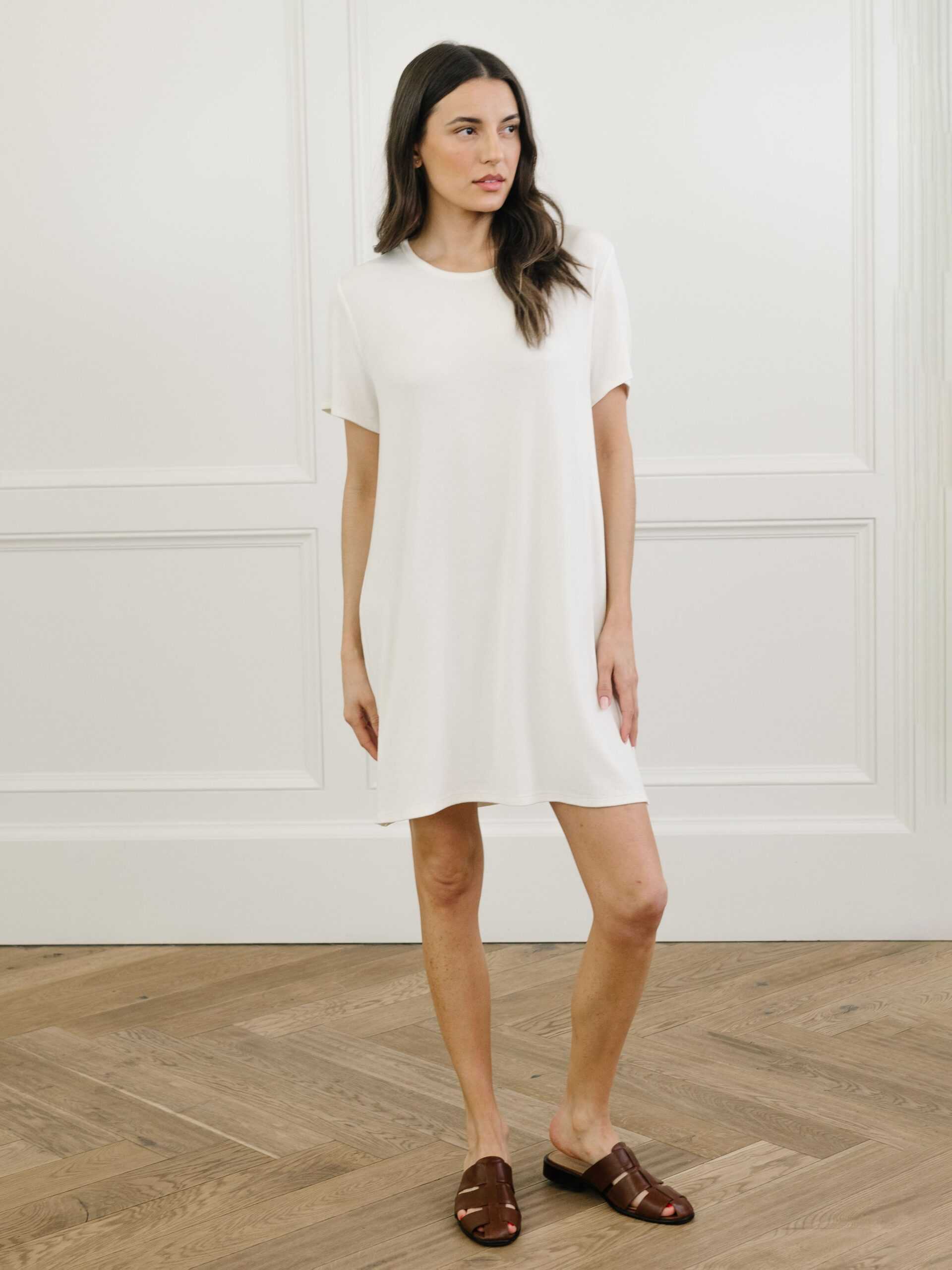 A woman standing in a white Cozy Earth bamboo rib-knit boyfriend sleep dress with short sleeves, knee-length, in a lounge or bedroom setting. |Color:Ivory