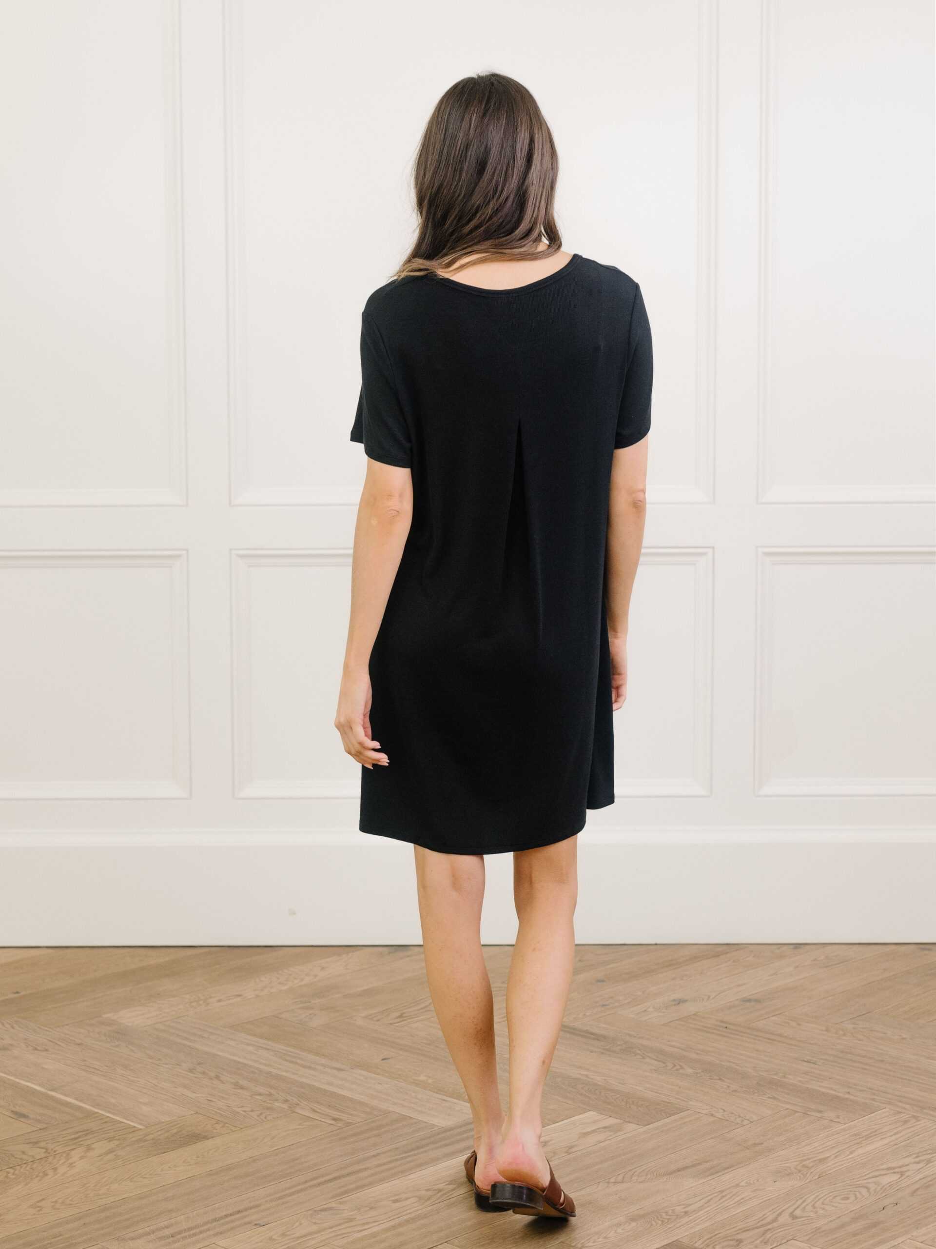 A woman facing backwards standing in a black Cozy Earth bamboo rib-knit boyfriend sleep dress with short sleeves, knee-length, in a lounge or bedroom setting. |Color:Black