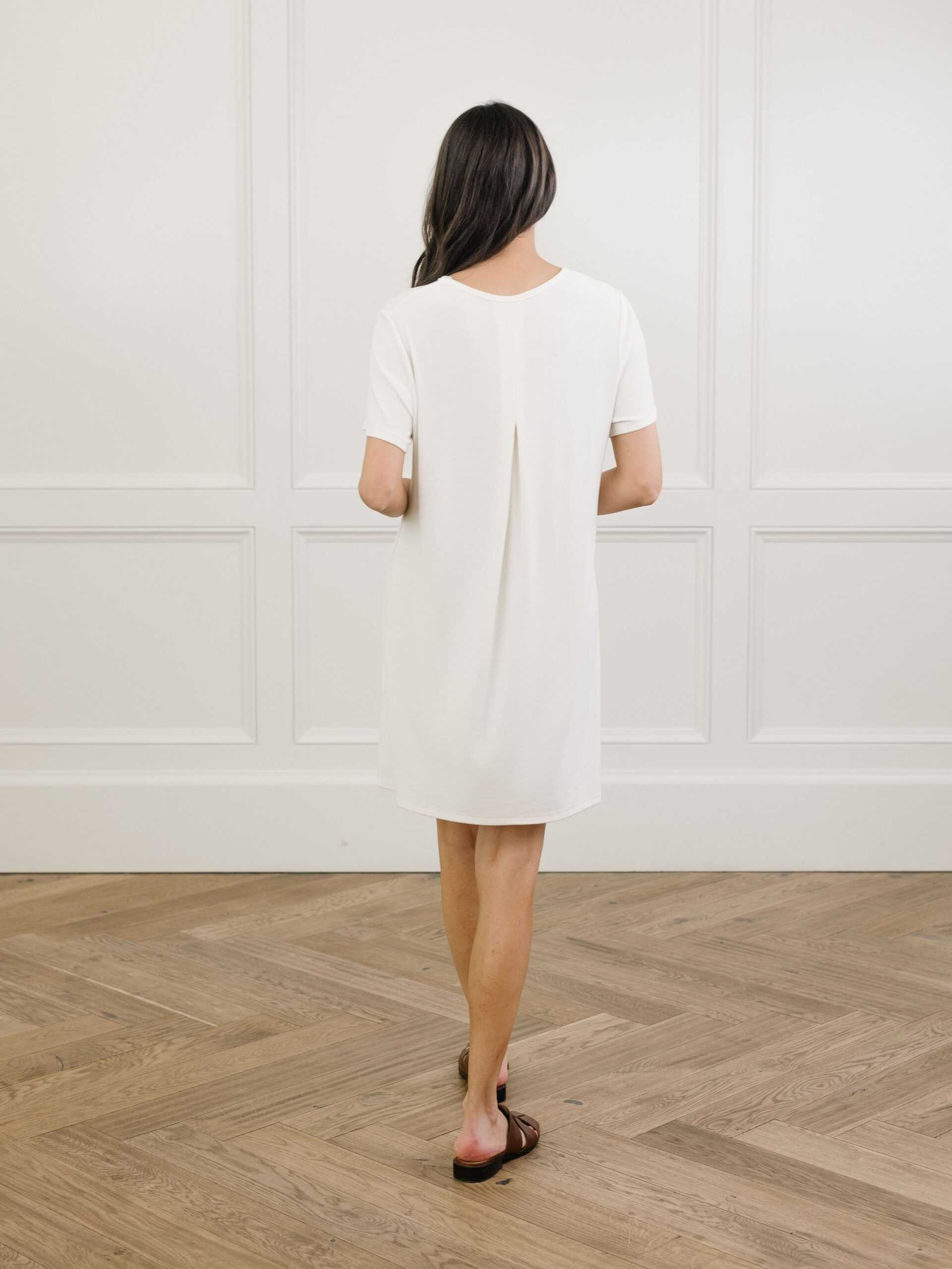 A facing backwards woman standing in a white Cozy Earth bamboo rib-knit boyfriend sleep dress with short sleeves, knee-length, in a lounge or bedroom setting. |Color:Ivory