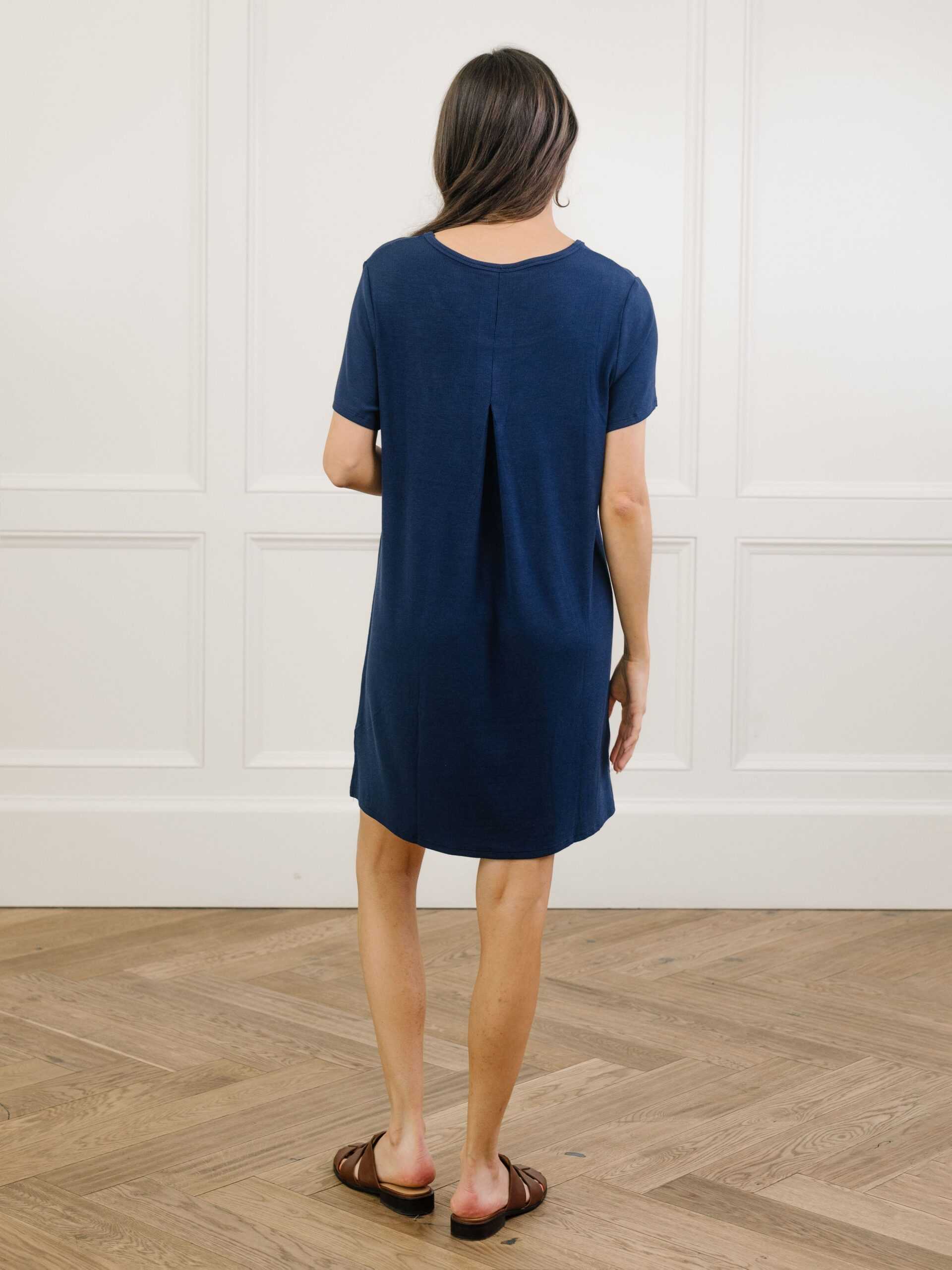 A woman facing backwards in a navy Cozy Earth bamboo rib-knit boyfriend sleep dress with short sleeves, knee-length, in a lounge or bedroom setting. |Color:Navy