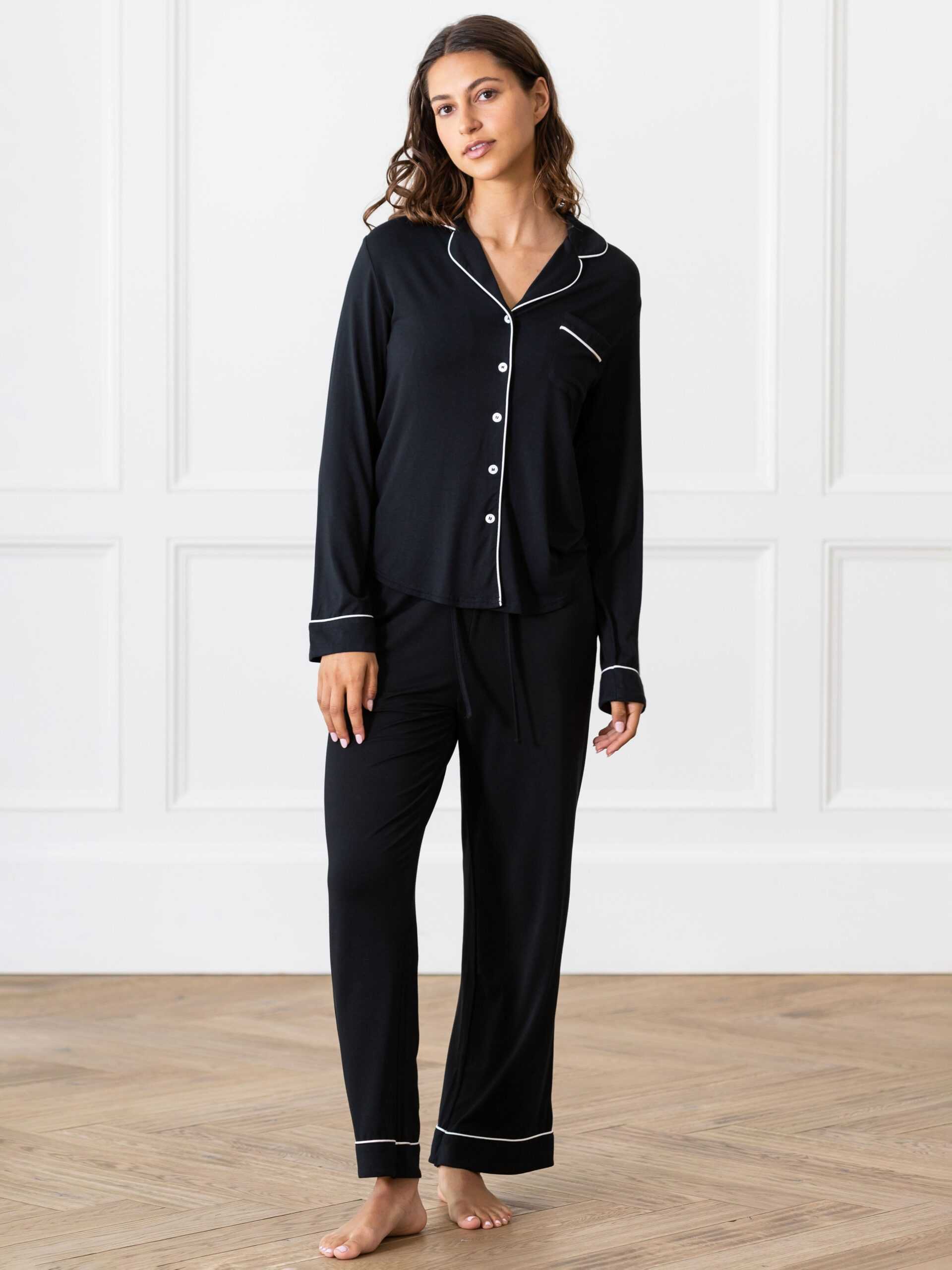 A woman stands indoors wearing the Women's Bedtime Bundle from Cozy Earth, a black pajama set with white piping that includes a long-sleeve button-up top and matching pants. She poses against a white paneled wall and wooden floor, looking at the camera with a neutral expression. |Color:Black