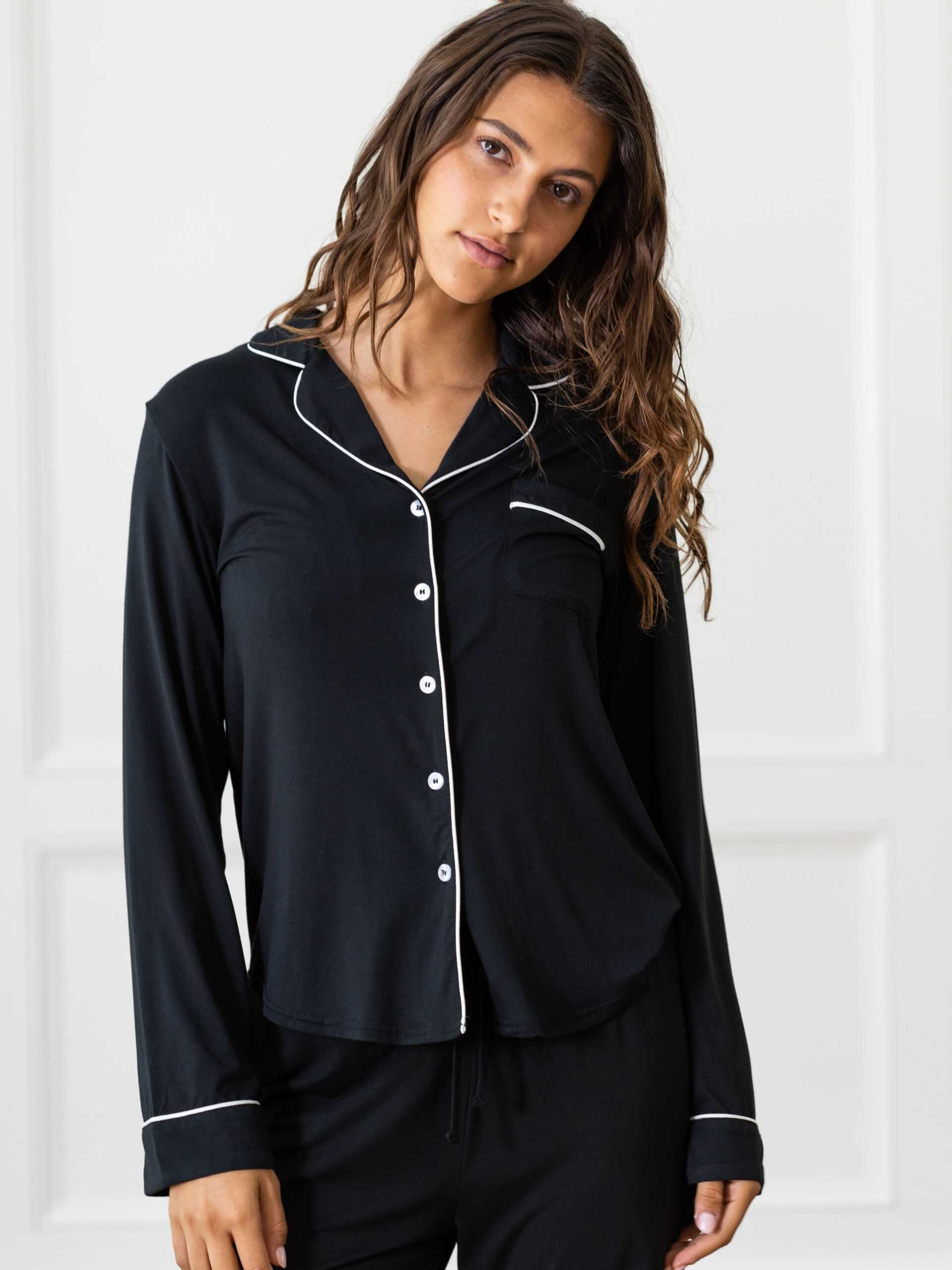 A person with long, wavy hair stands indoors wearing Cozy Earth's Women's Bedtime Bundle, featuring a black pajama top adorned with white piping along the collar, button placket, and cuffs. The top includes a chest pocket and is paired with matching black pajama pants. The background showcases a light-colored wall with decorative trim. |Color:Black