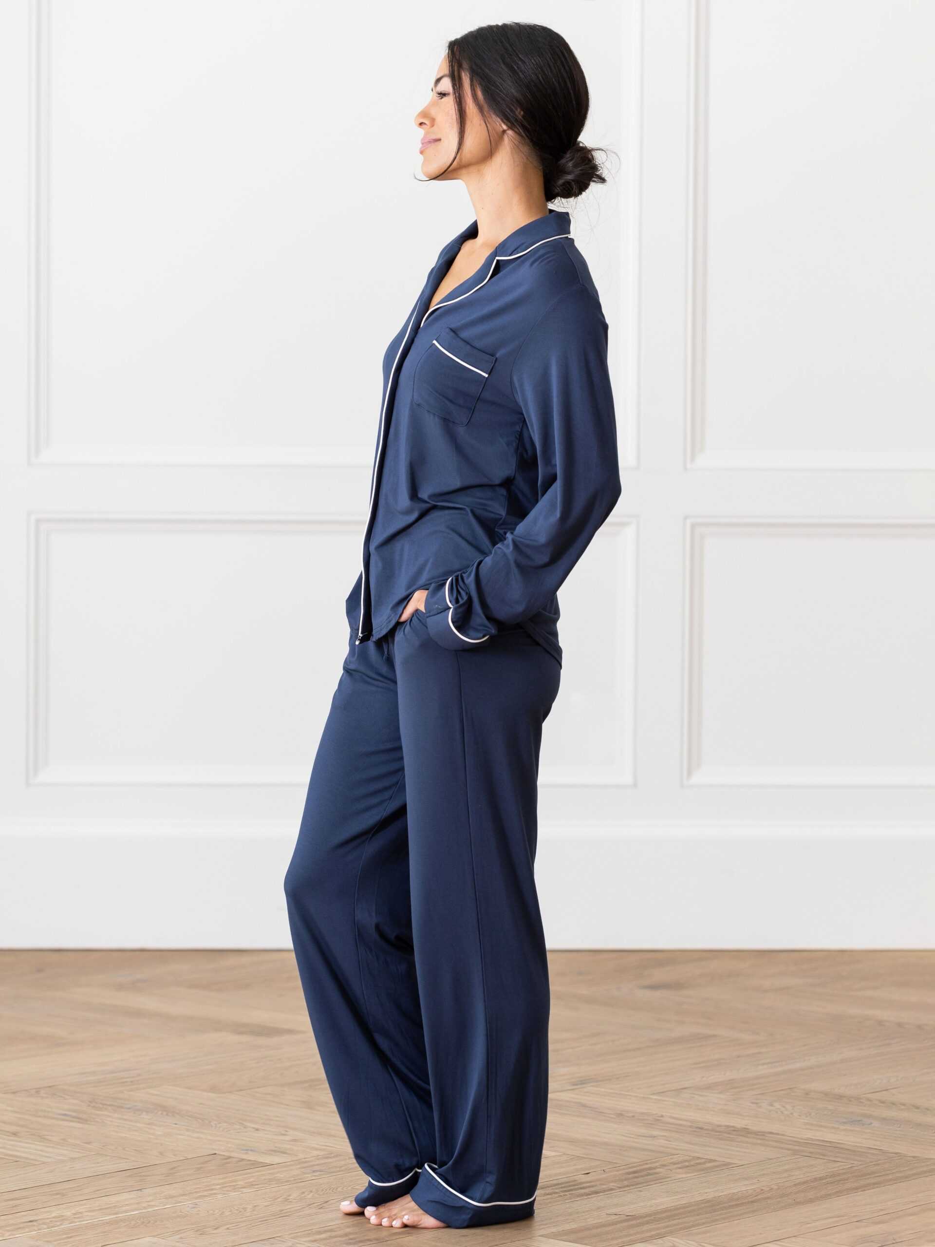 A person wearing the Women's Bedtime Bundle from Cozy Earth stands on a wooden floor with white paneled walls in the background. They have their hands in their pockets and are looking to the side. The dark blue, long-sleeve pajama set features white piping along the collar, cuffs, and pockets. |Color:Navy