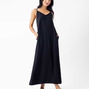 A person with long, dark hair wearing the Cozy Earth Women's Sunset Slip Dress stands against a plain white background. They wear black strappy sandals and have their hands in the dress's side pockets, smiling gently. |Color:Jet Black