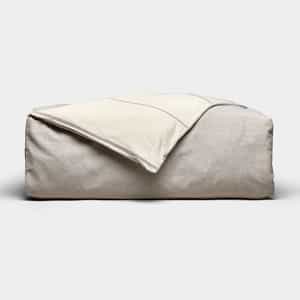 The Baja Duvet Cover by Cozy Earth, in light beige, is shown with a cream-colored blanket draped diagonally over it against a plain, light background.|Color:Coal Stripe/Striped Alabaster