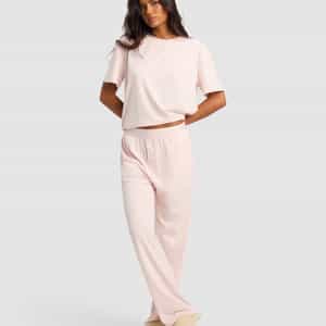A woman poses against a light gray background wearing the Cozy Earth Women's First Layer Pajama Short Sleeve Tee & Pant Set in light pink with beige slippers, her arms relaxed and legs crossed.|Color:Ballet